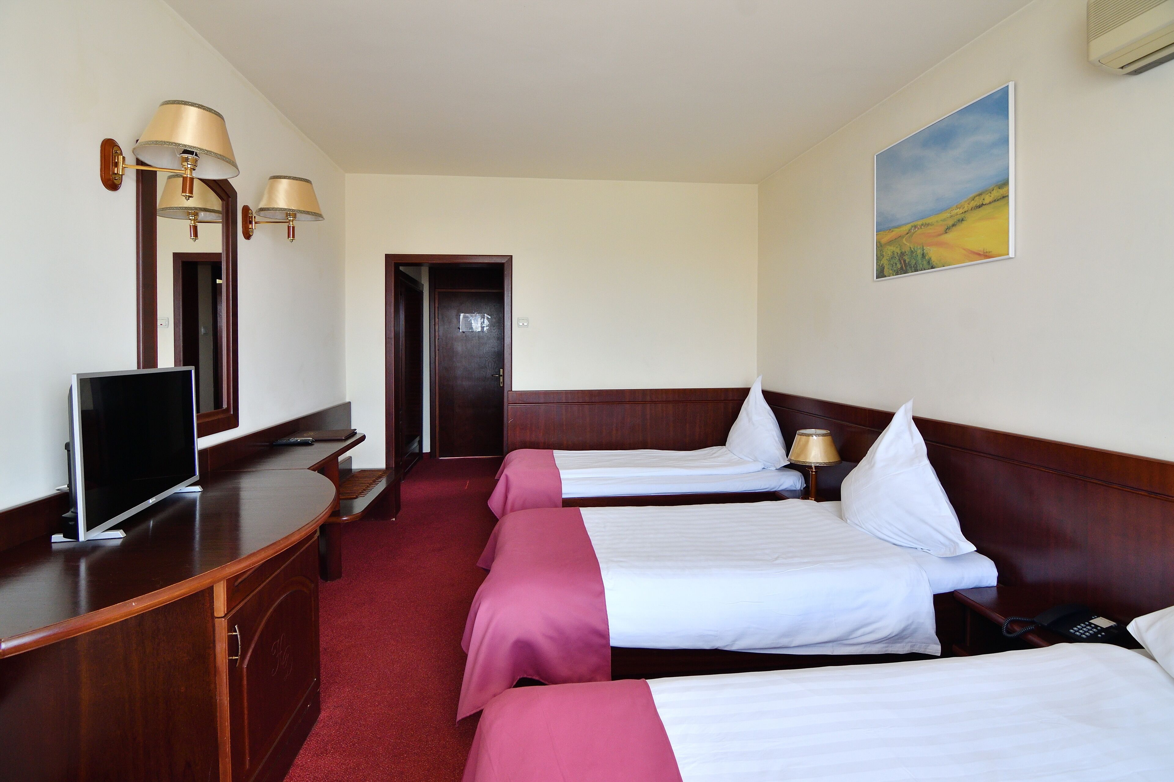 A hotel room with three beds, a desk with a flat-screen TV, and a painting on the wall.
