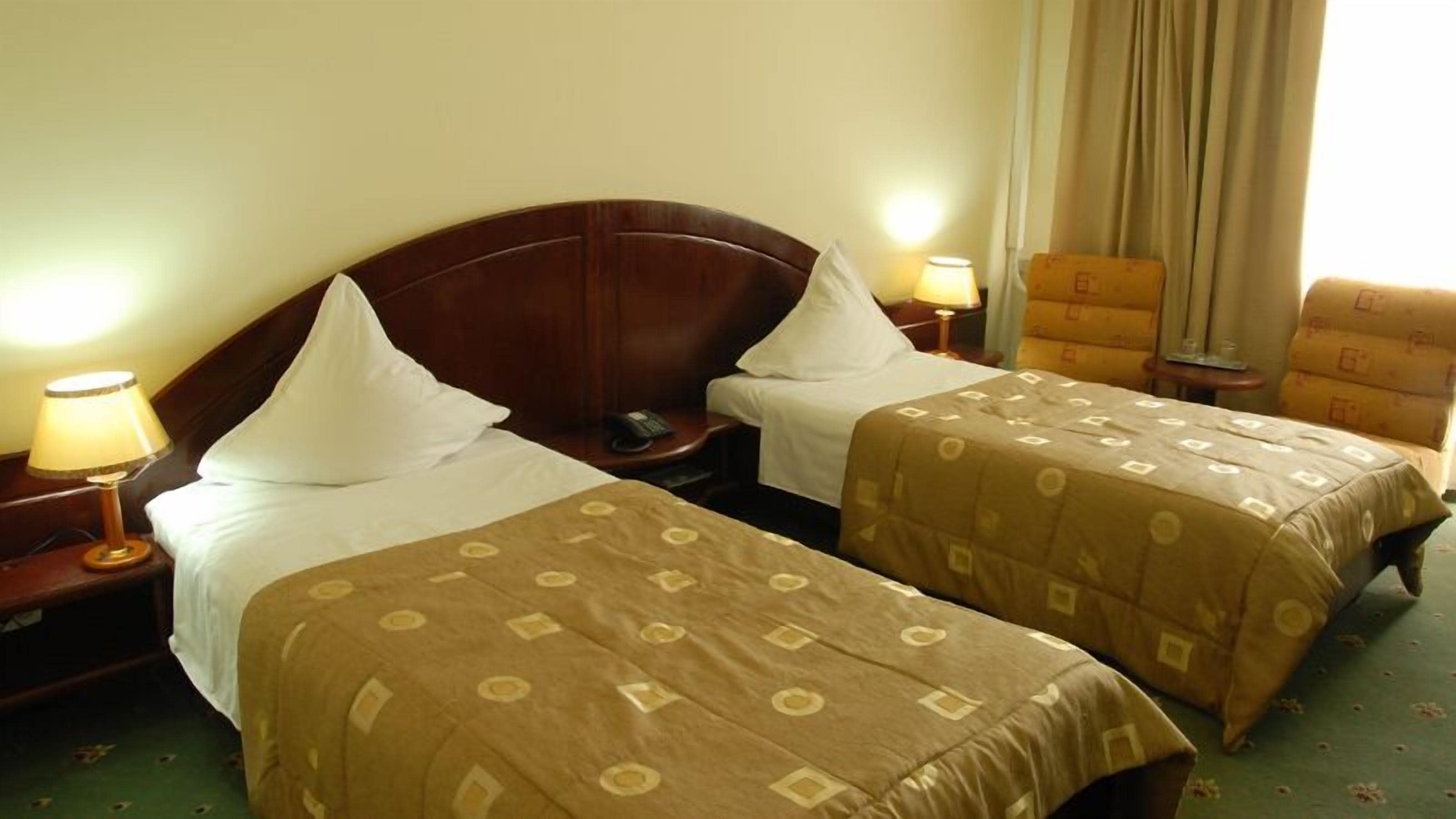 Een hotelkamer met twee bedden, elk met een houten hoofdbord en witte kussens. Er zijn nachtkastjes met lampen en een klein tafeltje met een telefoon. De kamer heeft een groen vloerkleed en beige gordijnen.