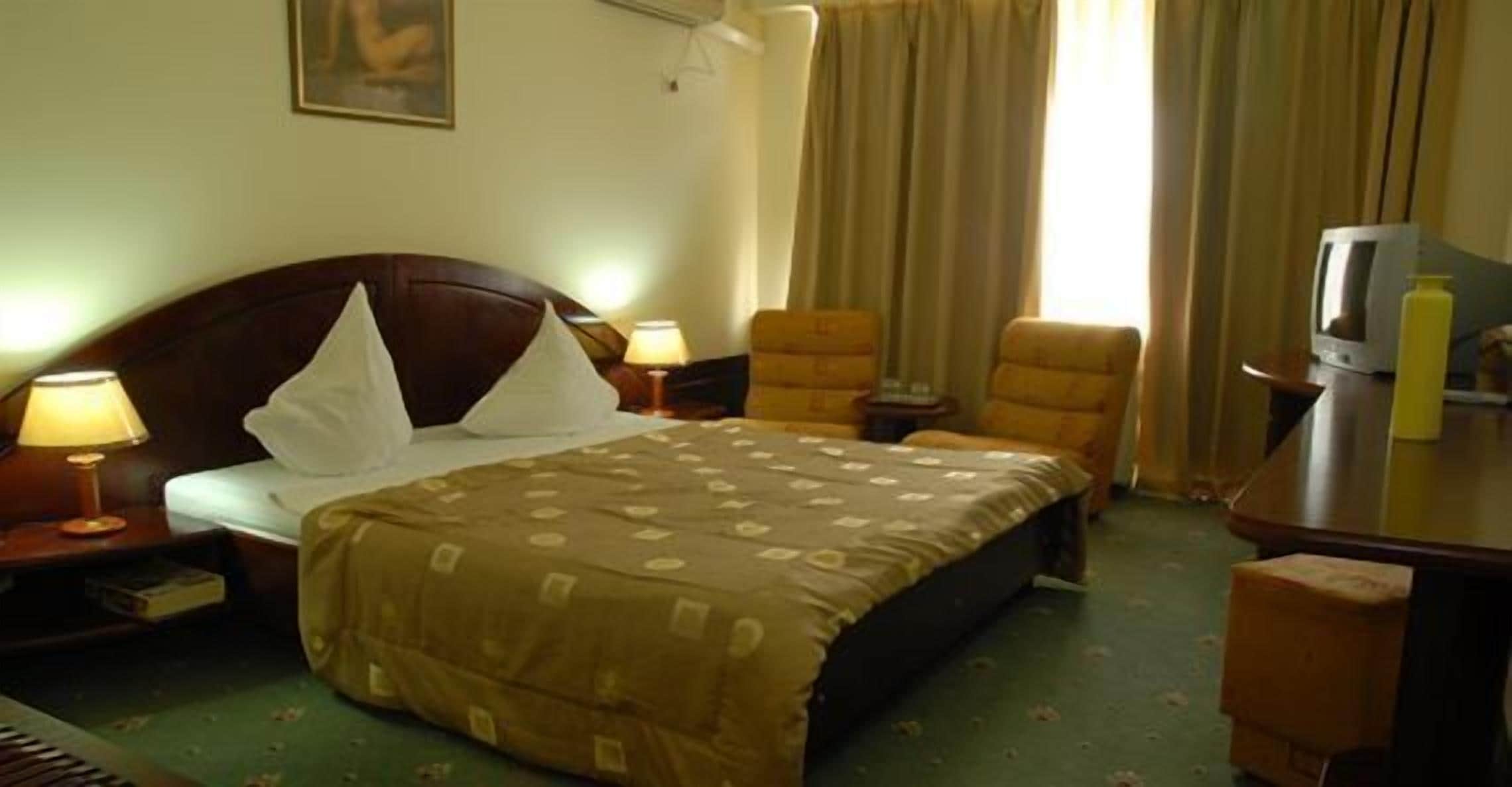 A hotel room with a bed, two bedside lamps, a television, a desk with a lamp, and a chair.