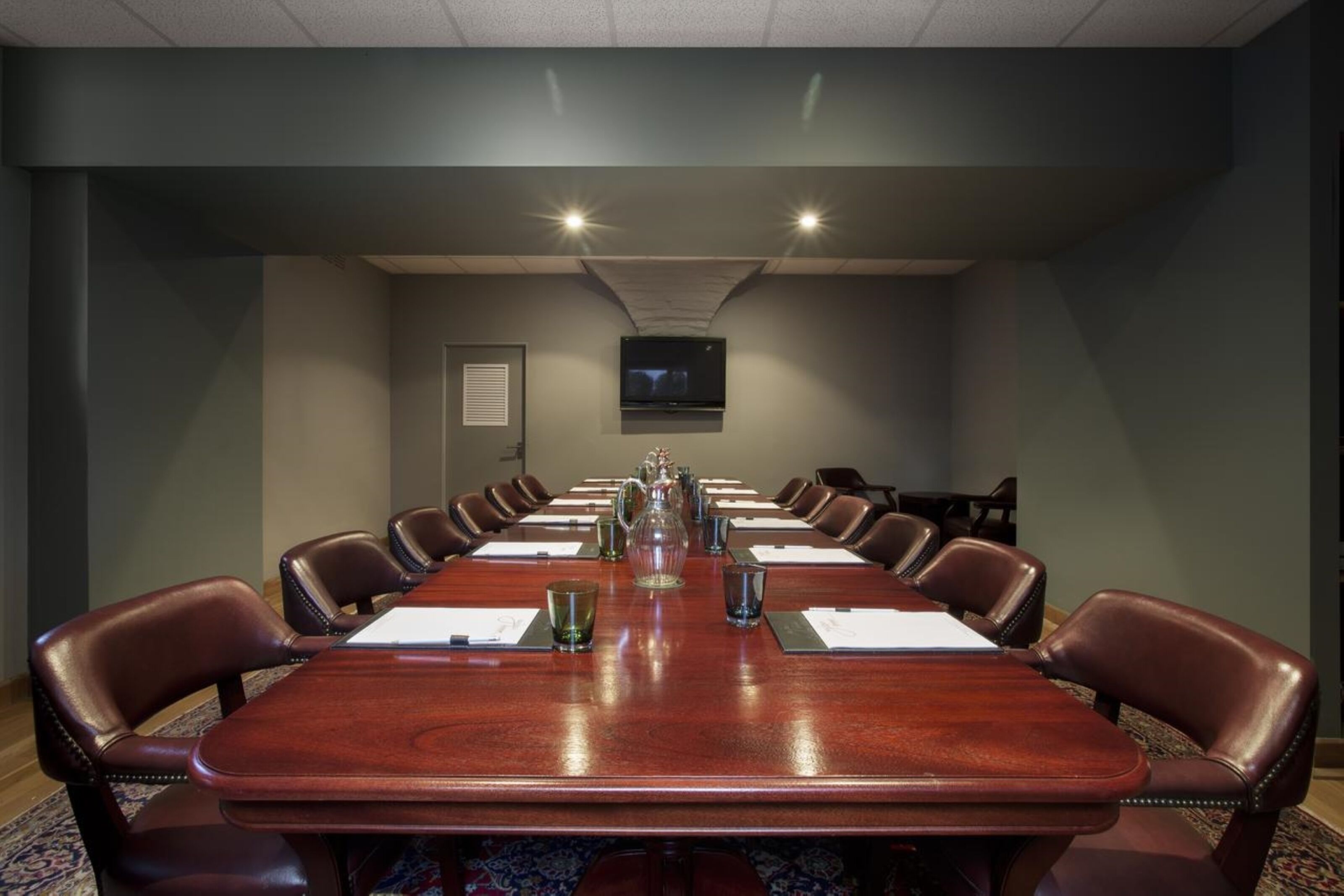 A conference room with a long wooden table, leather chairs, and a flat-screen TV mounted on the wall.