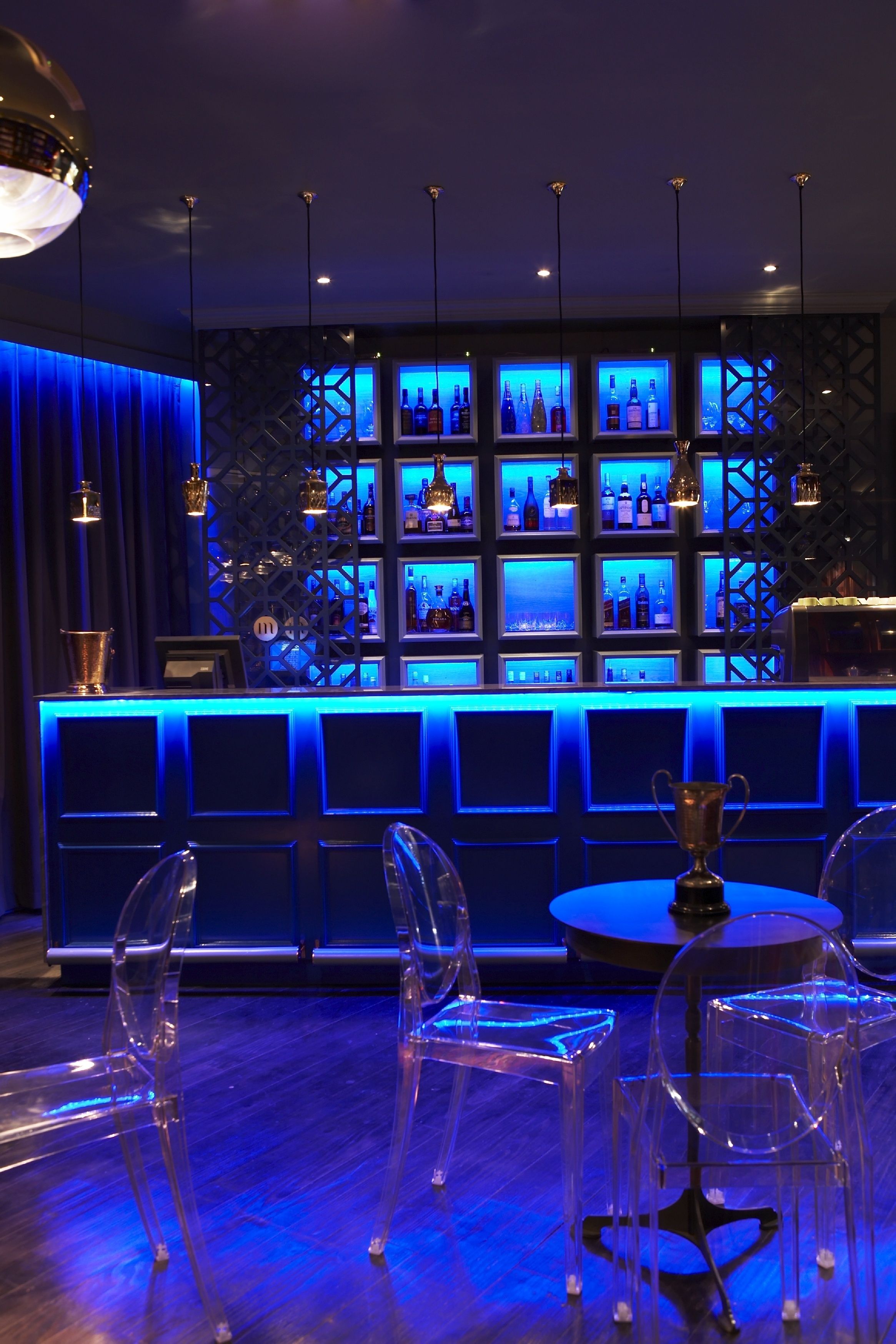 A bar area with a blue-lit bar counter, clear chairs, and a trophy on a round table.