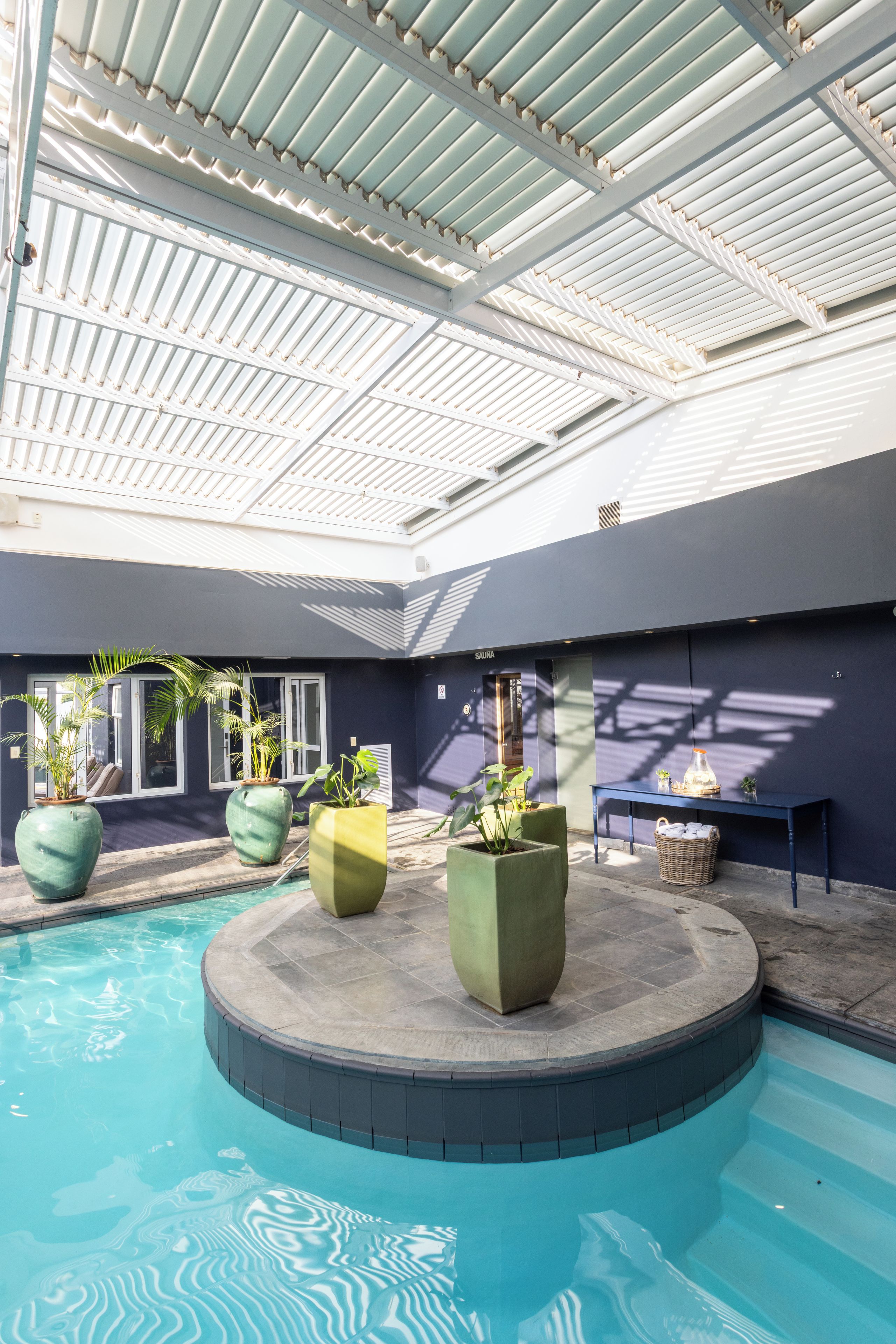 An indoor swimming pool area with a covered roof, seating around the pool, and potted plants.