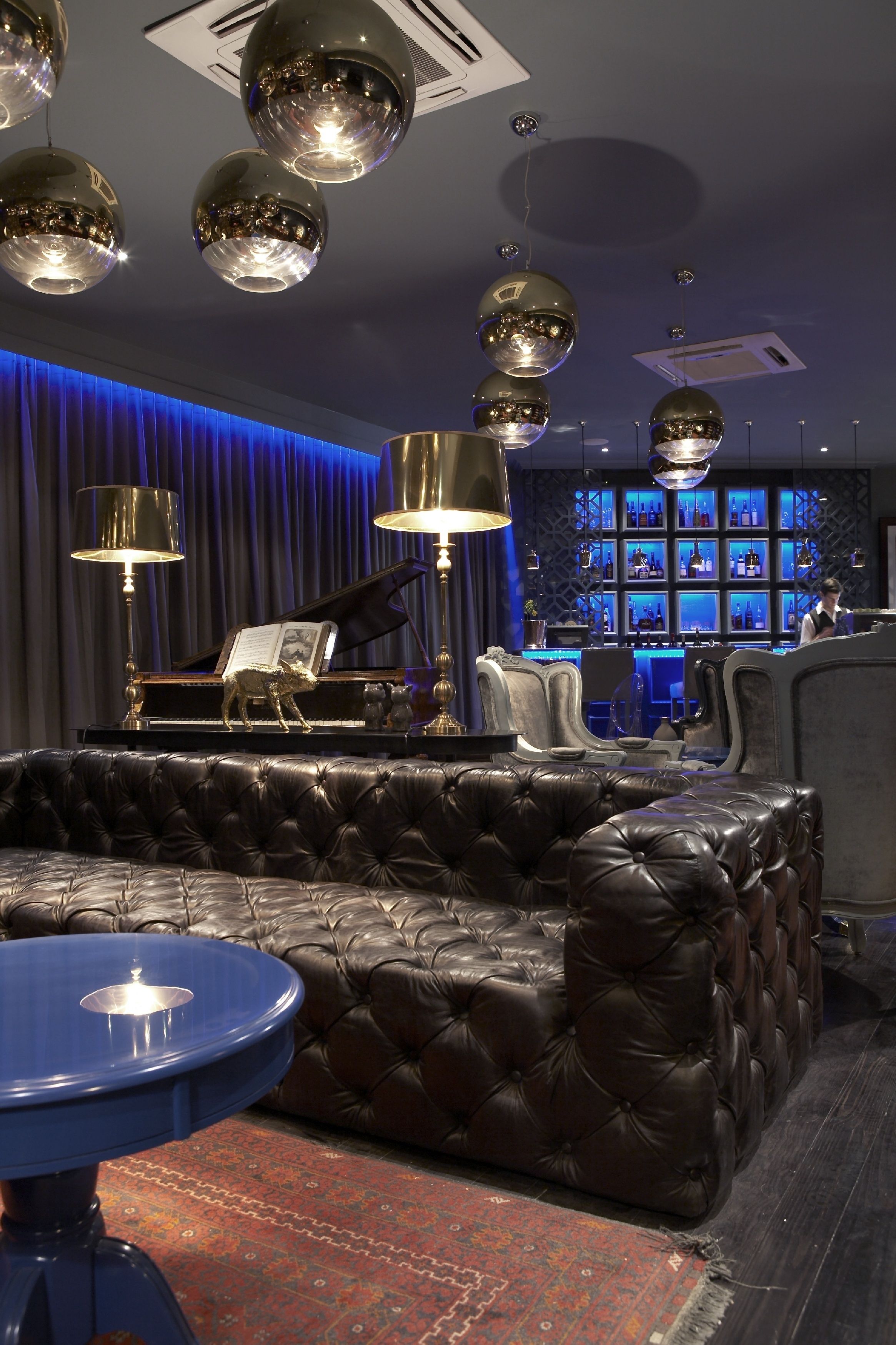 A luxurious lounge area with a grand piano, plush leather sofas, and a bar with blue lighting.