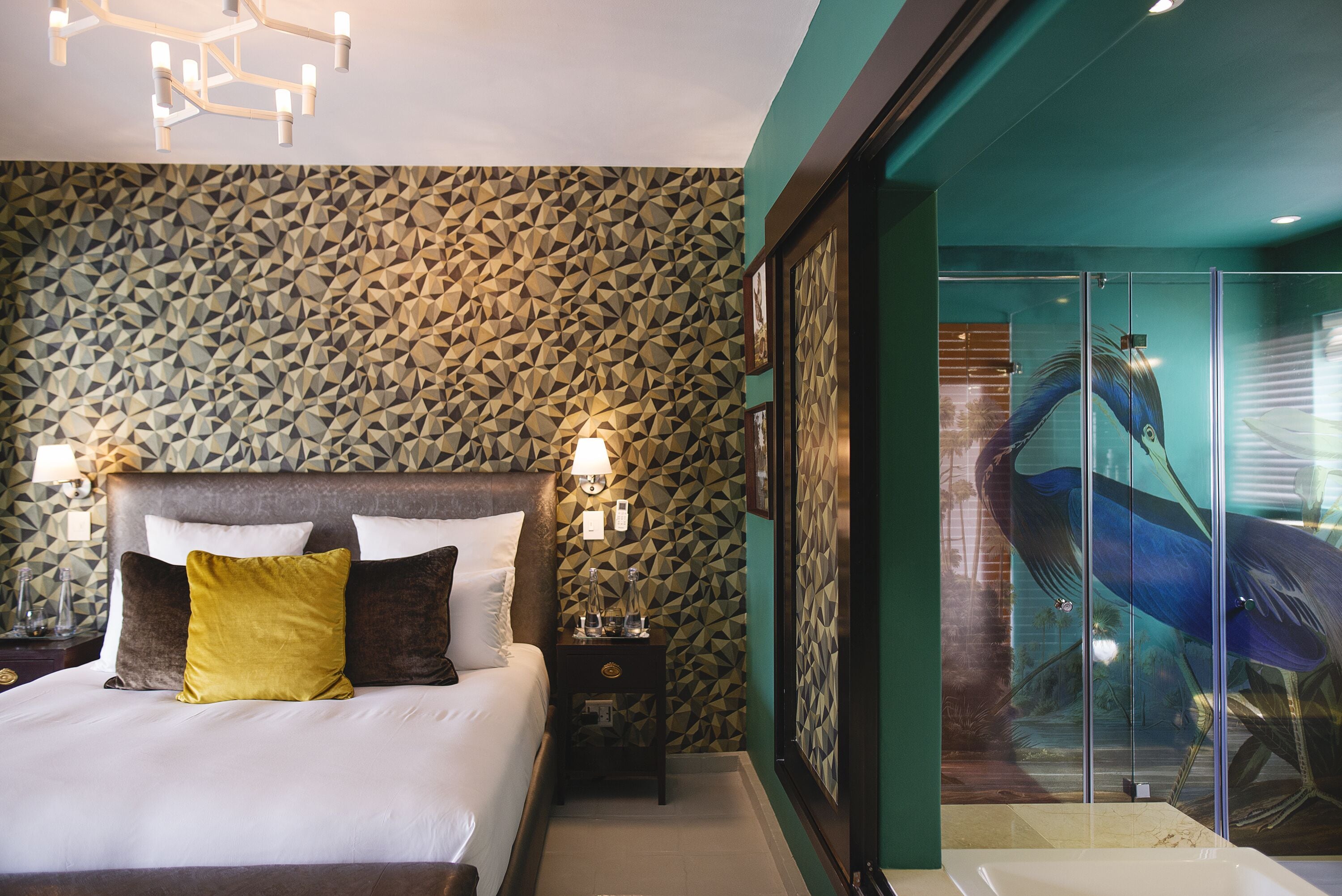 Ein Schlafzimmer mit einem großen Bett, einem Nachttisch, einer Wand mit geometrischem Muster und einer Glastür zu einem Badezimmer mit einem großen Spiegel und einem blauen Kunstwerk.