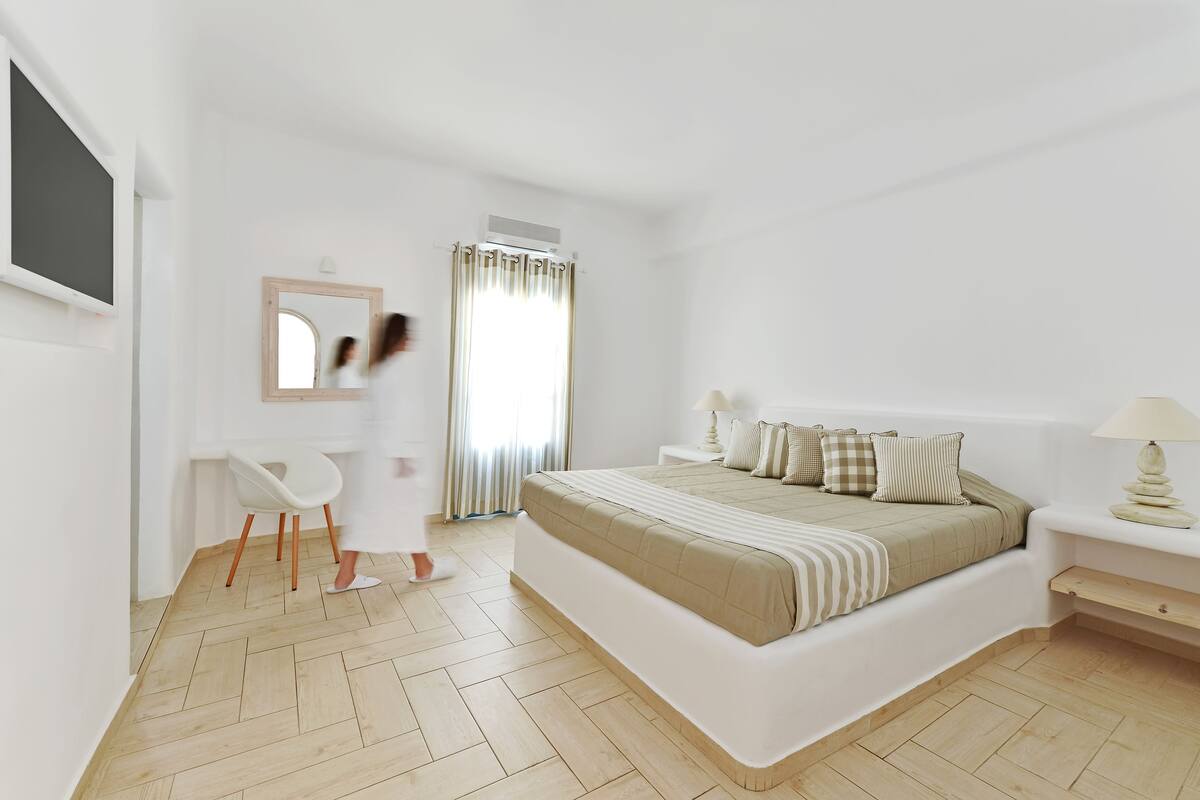 Un dormitorio moderno con una cama grande, una mesita de noche con lámpara, una silla, un escritorio con espejo y una ventana con cortinas.