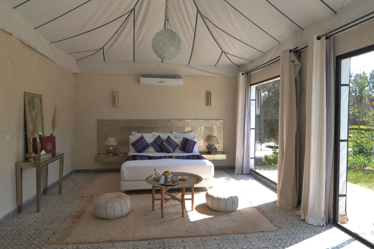 A spacious bedroom with a large bed, bedside tables, a round coffee table, and a view of greenery through a large window.