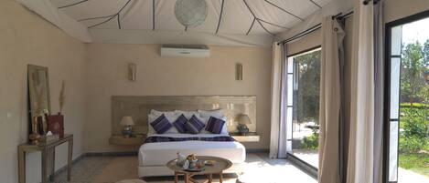 A spacious bedroom with a large bed, bedside tables, a round coffee table, and a view of greenery through a large window.
