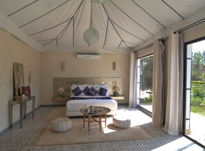A spacious bedroom with a large bed, bedside tables, a round coffee table, and a view of greenery through a large window.