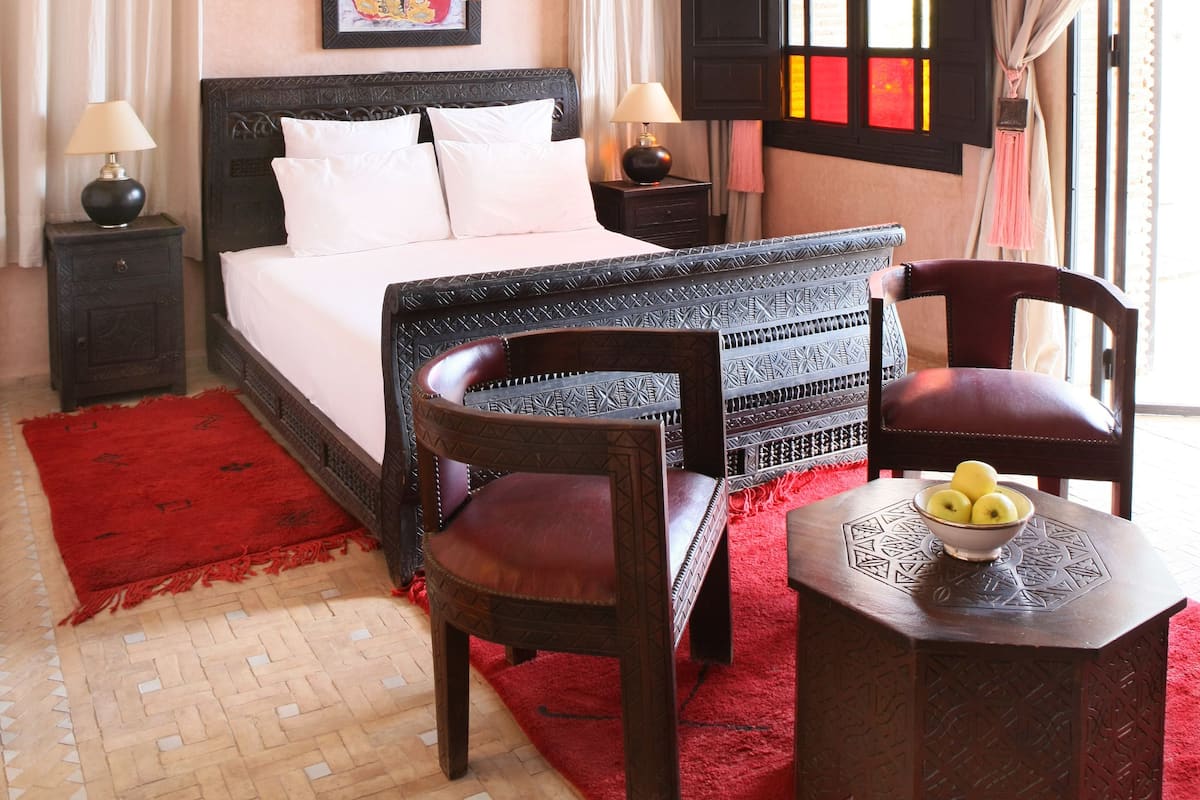 A bedroom with a bed, two nightstands, a chair, a small table with fruit, a red rug, a patterned floor, a hanging light fixture, and a window with curtains.