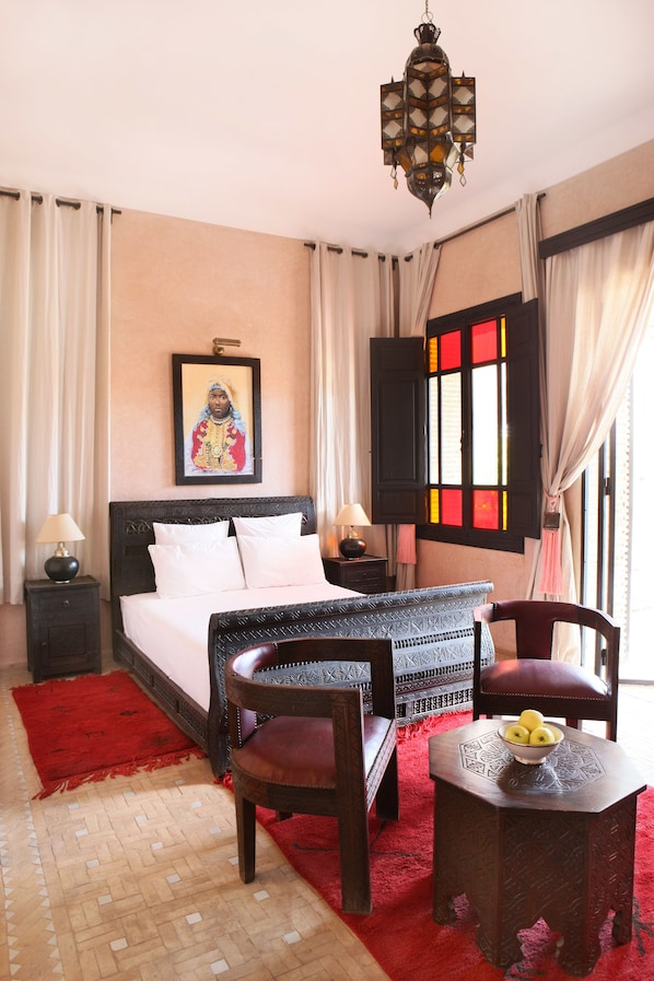 Une chambre à coucher comprenant un lit, deux tables de chevet, une chaise, une petite table avec des fruits, un tapis rouge, un sol à motifs, un luminaire suspendu et une fenêtre avec des rideaux.
