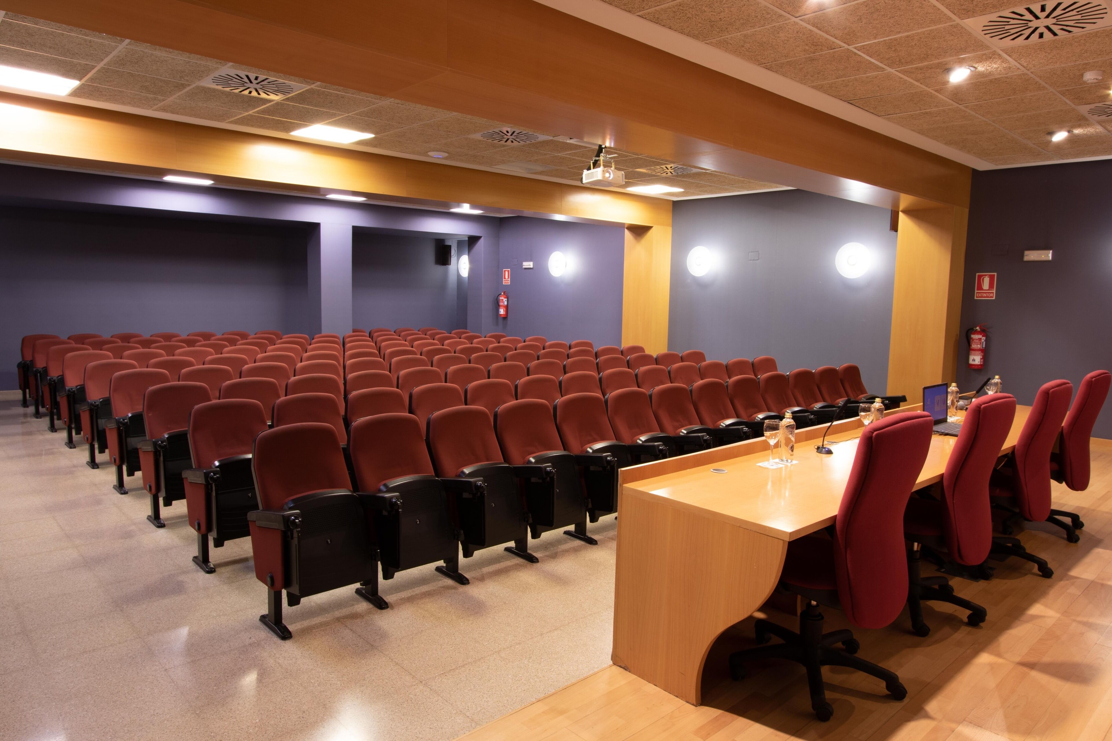 Un salón de conferencias con filas de sillas, una mesa larga y una pantalla para proyector.