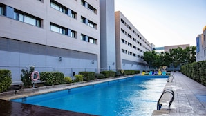 A swimming pool with a wooden deck, surrounded by a modern building with multiple windows and a clear sky.