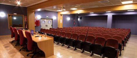 Un salón de conferencias con filas de sillas rojas, una mesa de madera con computadoras portátiles y una pantalla de proyector.