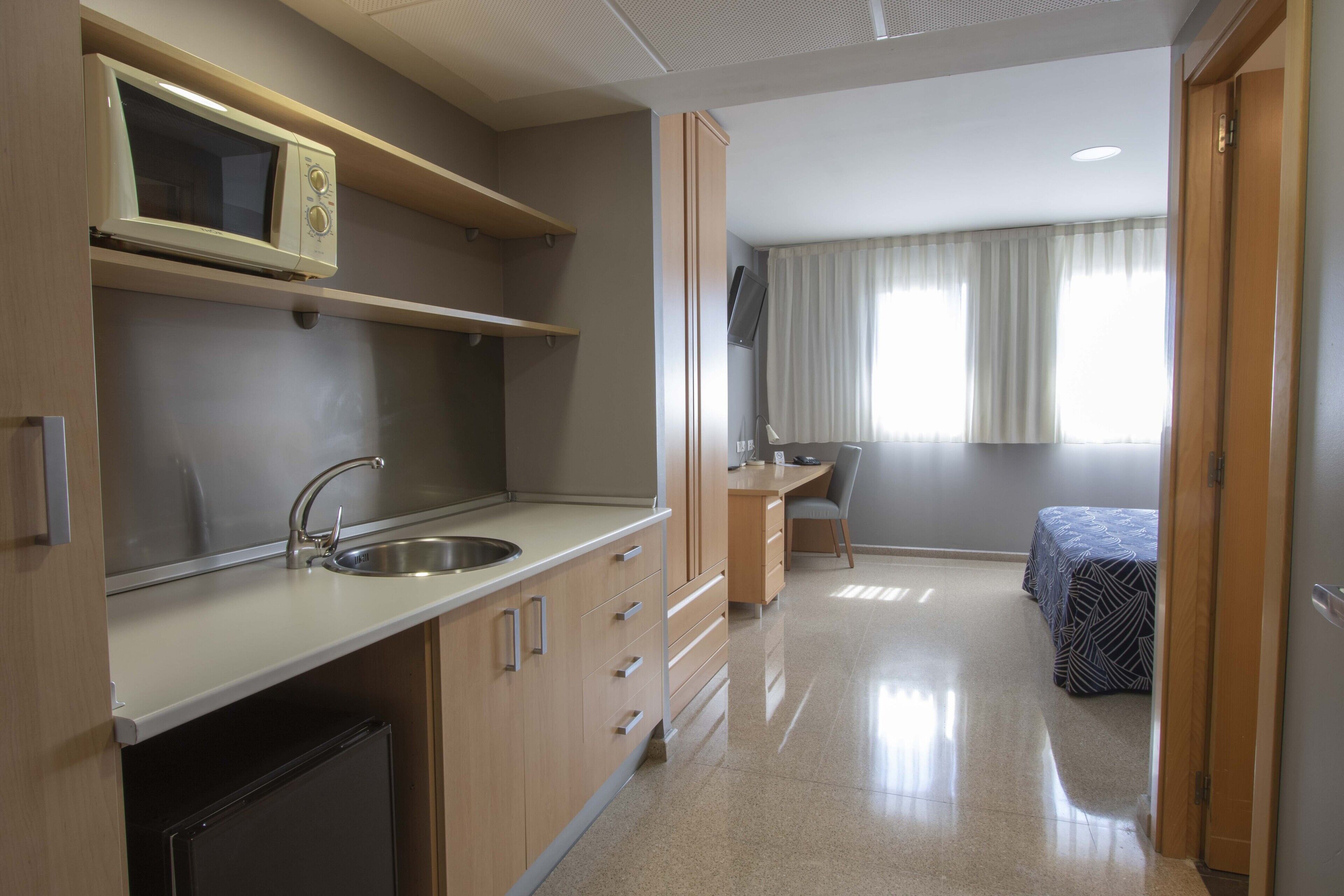 Cocina compacta con microondas, lavamanos y armarios. Incluye un refrigerador pequeño, un escritorio con silla y una cama con colcha estampada.