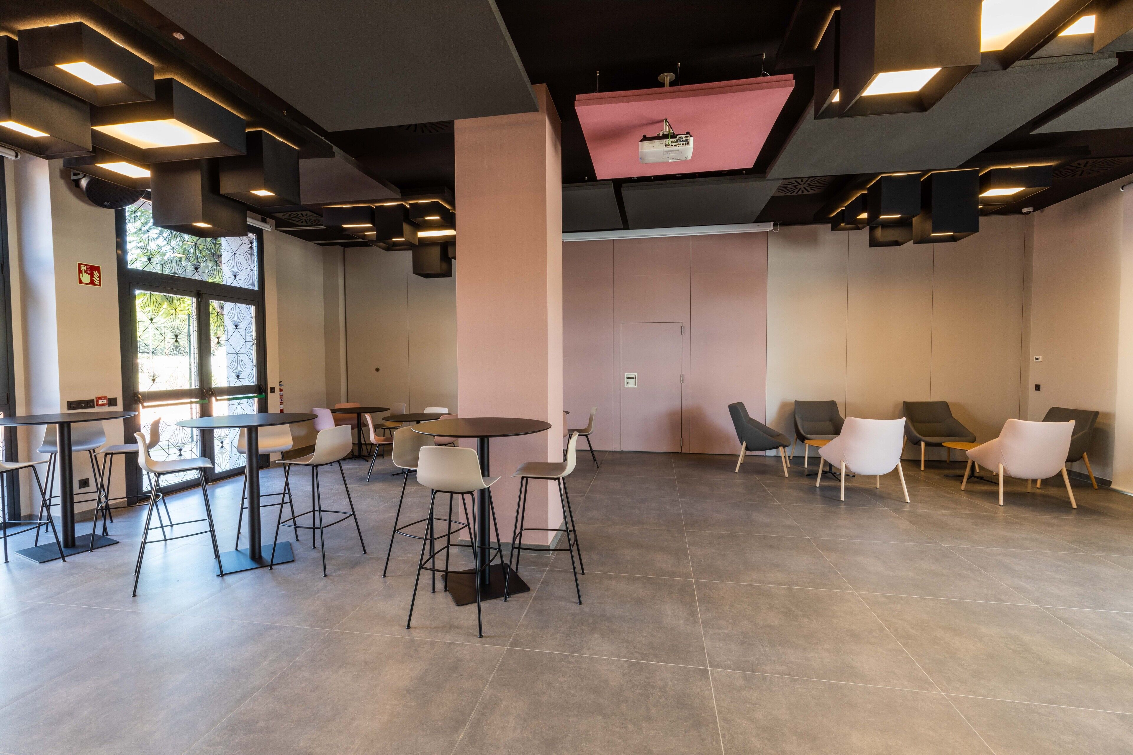 A modern indoor space with tables and chairs, a projector on the ceiling, and a door in the center.