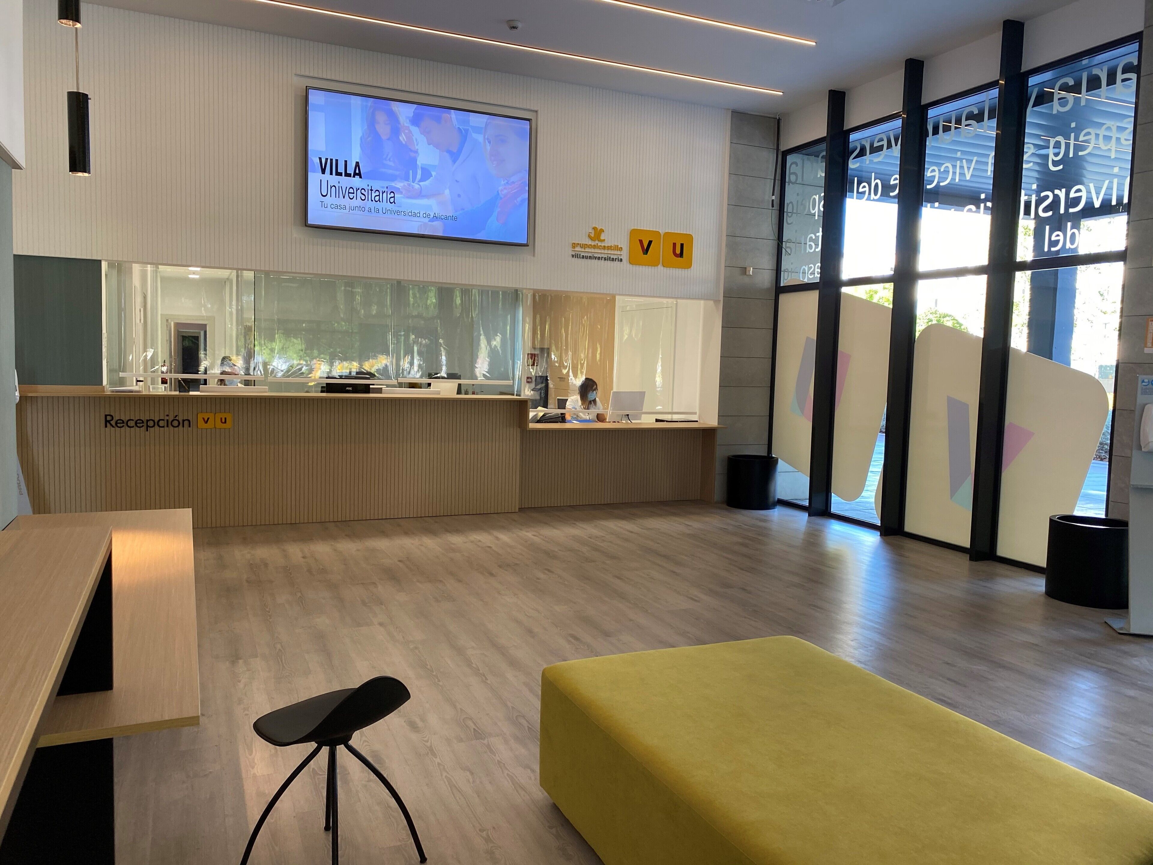 Moderni vastaanottoalue, jossa on puinen vastaanottotiski, suuri näyttö, jossa esitetään tekstiä »Villa Universidad de Alicante», ja keltainen oleskelualue.
