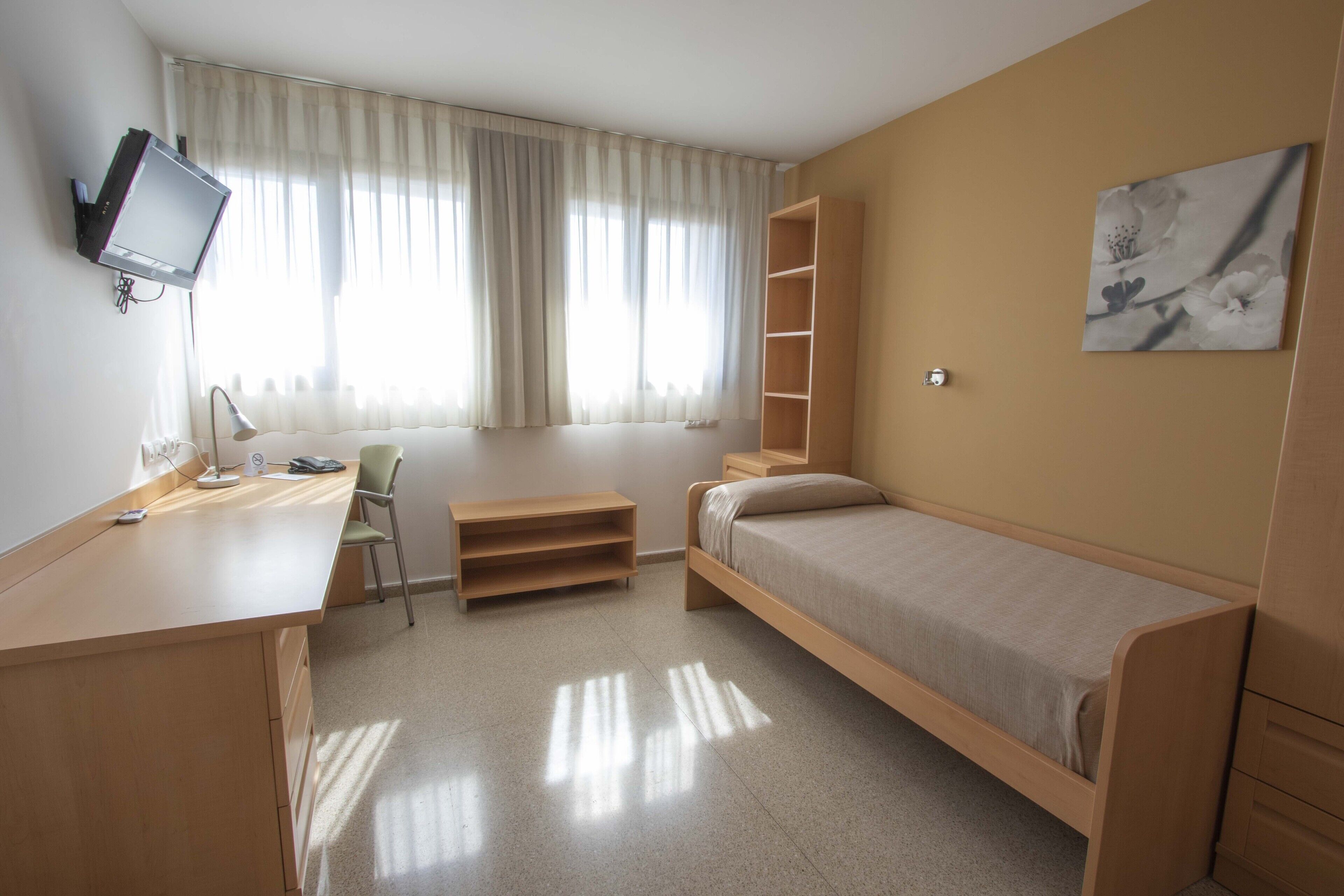 Una habitación pequeña y bien iluminada con escritorio, silla, cama y televisor montado en la pared.