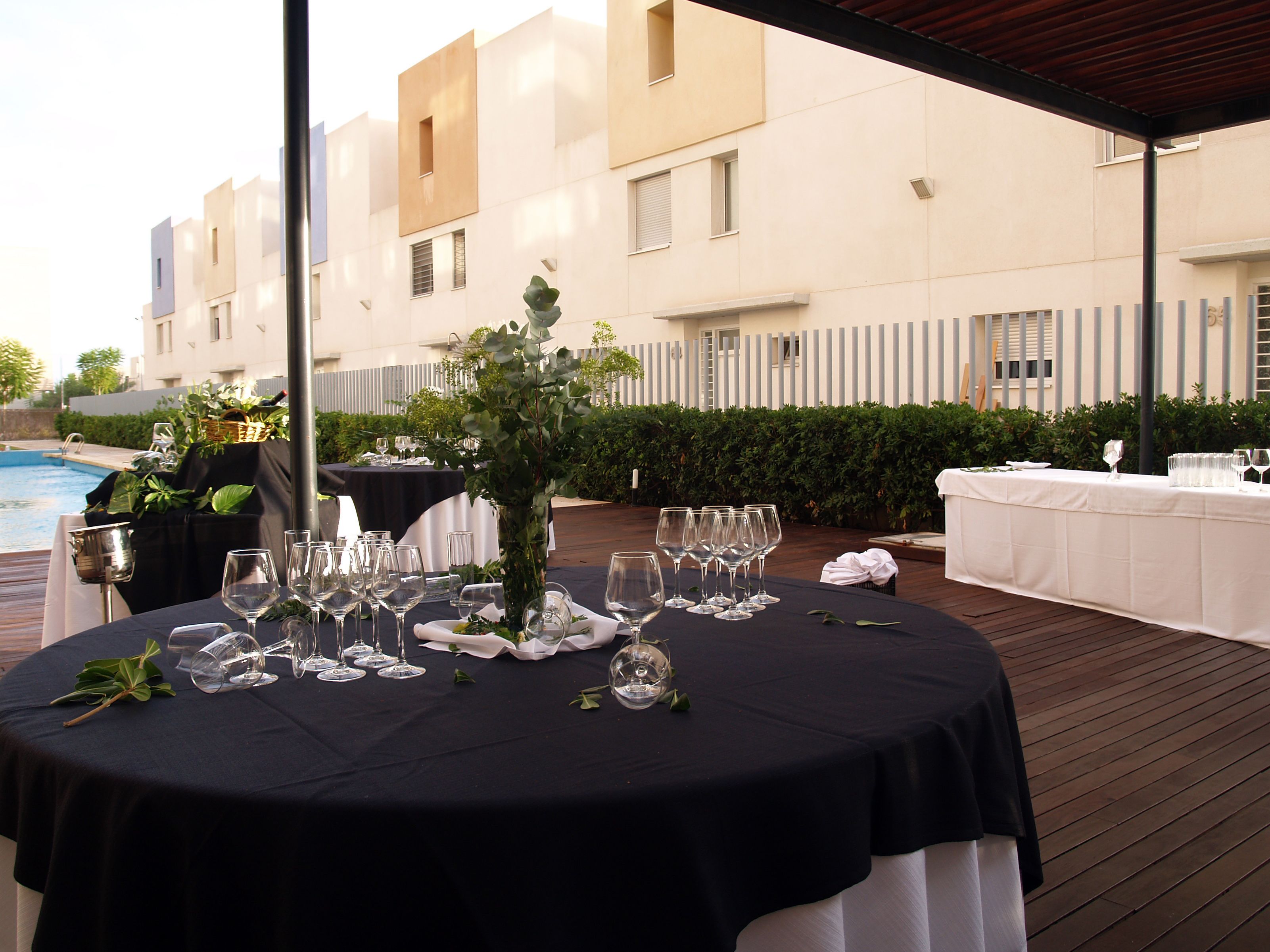Un área de comedor al aire libre con mesas redondas dispuestas para una comida, una piscina al fondo y un edificio con balcones.