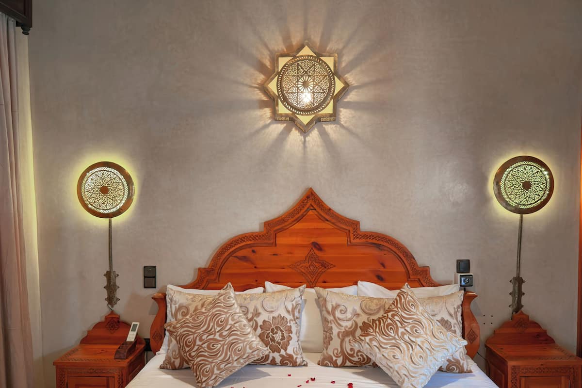 Una habitación de hotel con cabecera de madera, dos mesitas de noche con lámparas, cama con almohadas estampadas y una lámpara de pared decorativa.