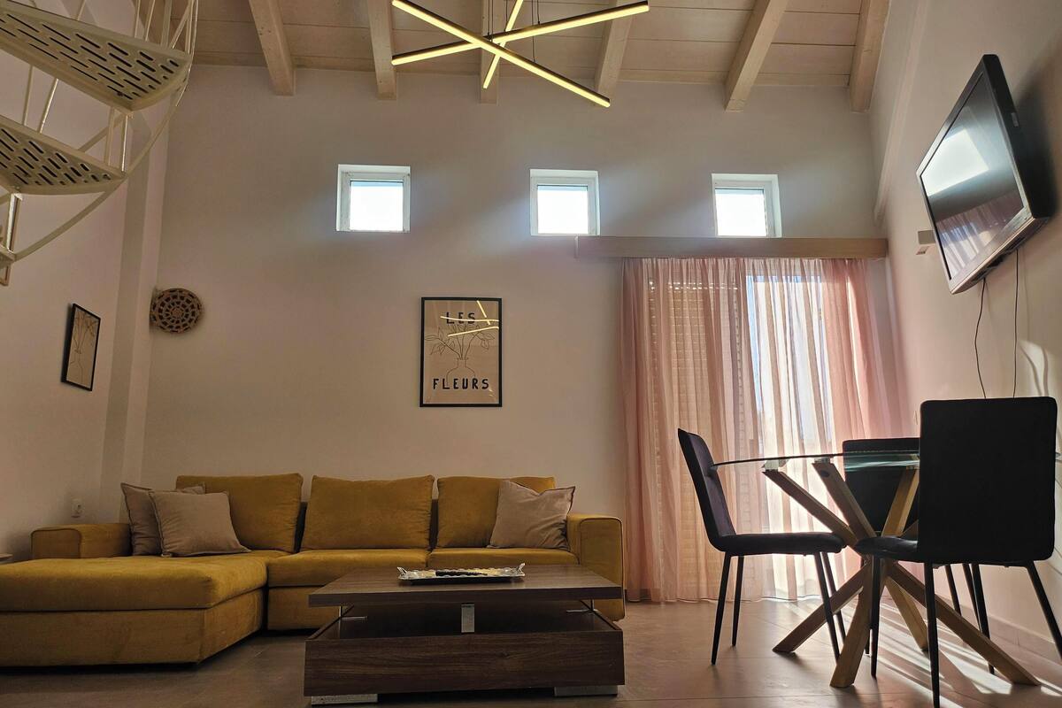 A modern living room with a sofa, coffee table, and a TV mounted on the wall. There are two windows with curtains, a ceiling light fixture, and a framed picture on the wall.