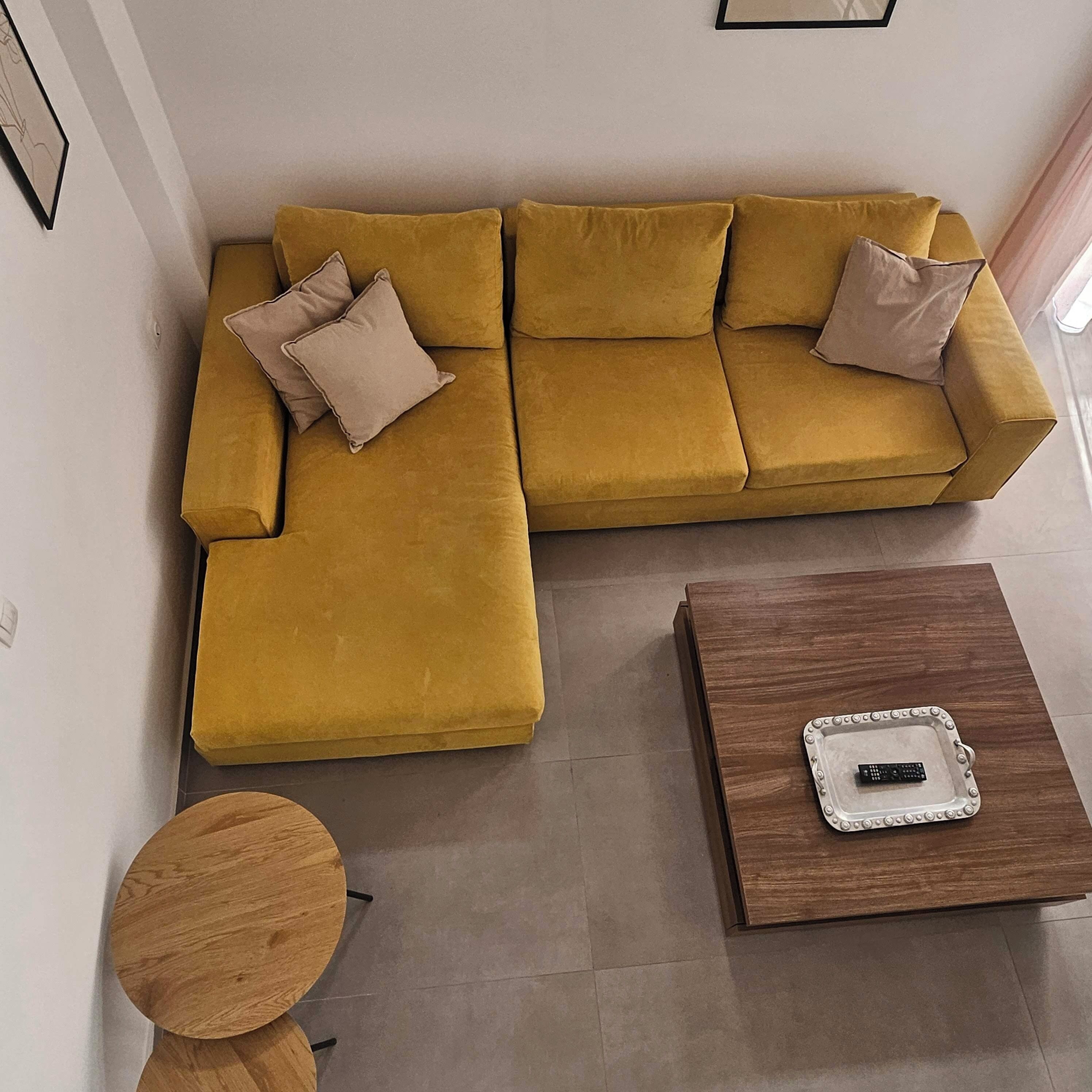 A living room with a mustard-colored sofa, two beige cushions, a wooden coffee table with a remote control, and a small round table.