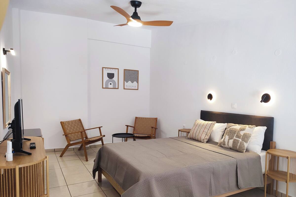 A bedroom with a bed, two chairs, a desk with a TV, a ceiling fan, and wall-mounted lights.