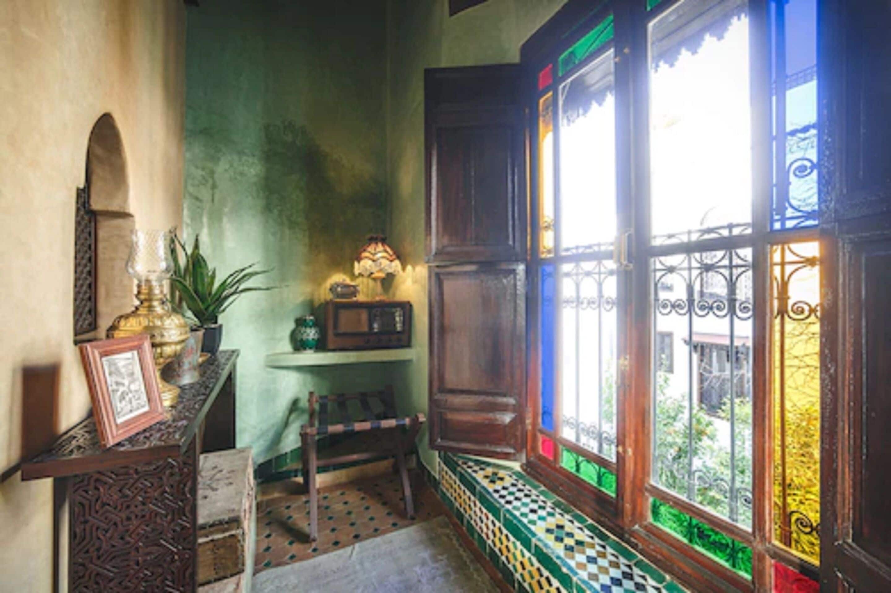 A room with a wooden bench, a small table with a lamp, and a window with colorful stained glass.