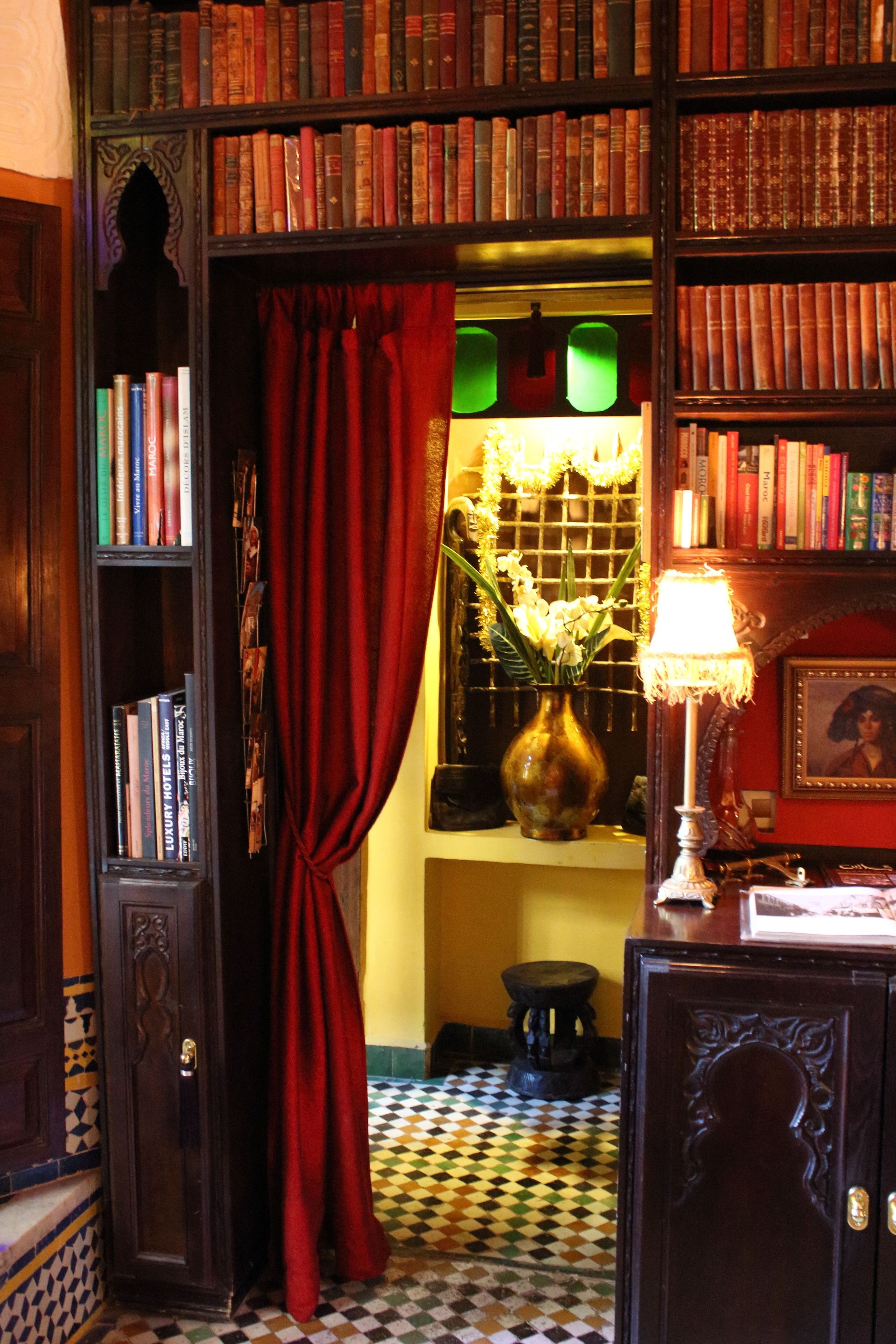 Um quarto com chão quadriculado, uma estante cheia de livros, uma cortina vermelha e um abajur.