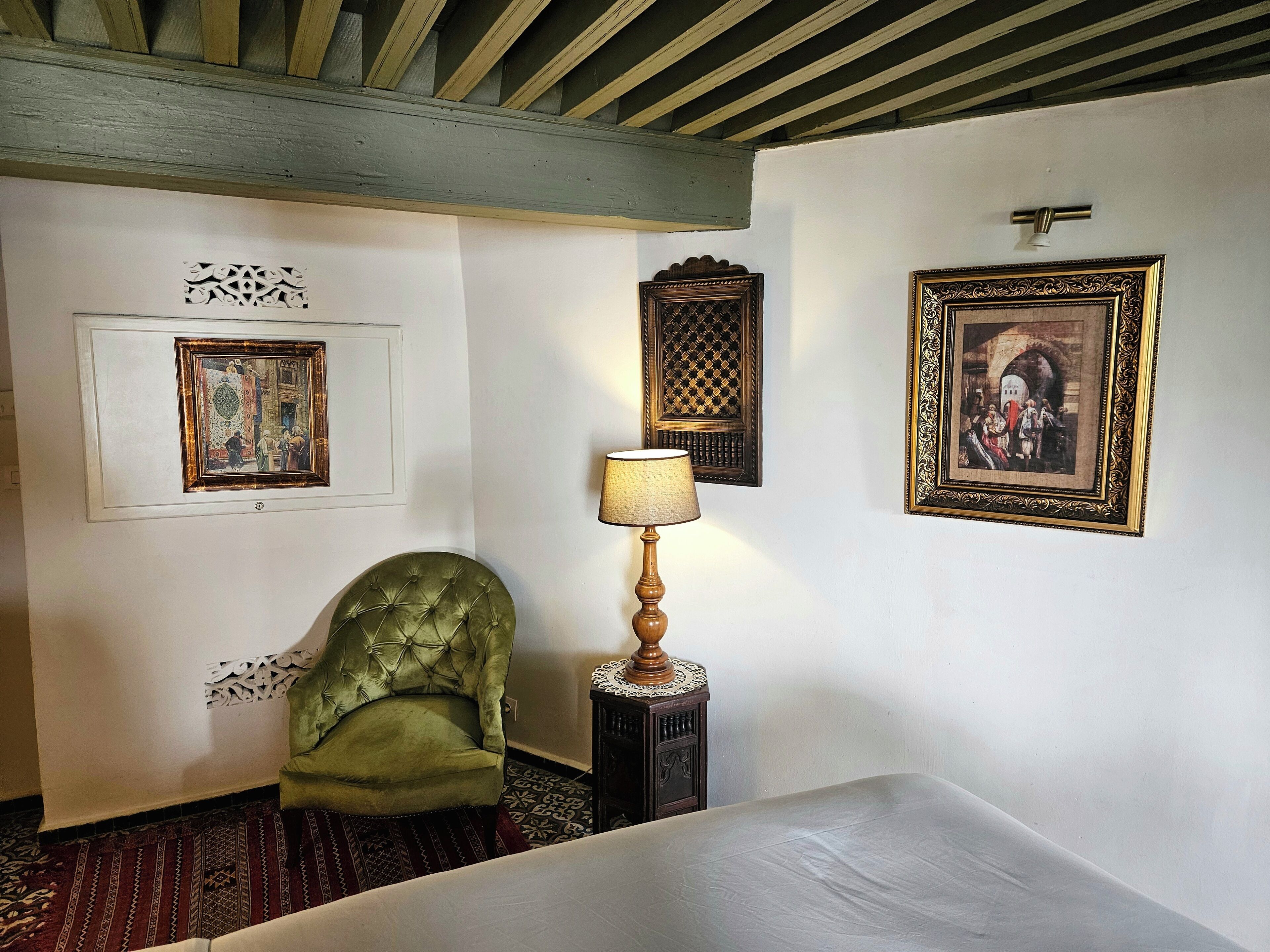 A hotel room with a green upholstered chair, a bedside table with a lamp, and three framed artworks on the walls.