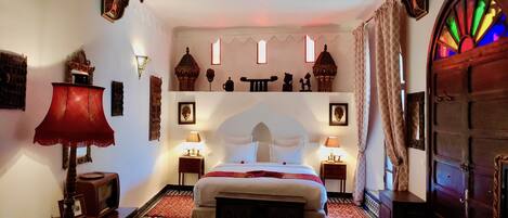A traditional room with a bed, two bedside tables with lamps, a red sofa, patterned rugs, and a decorative wall with shelves.