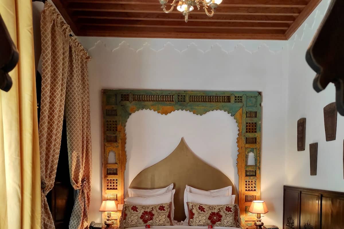 A bedroom with a large bed, a chandelier, patterned curtains, and a decorative wall headboard.