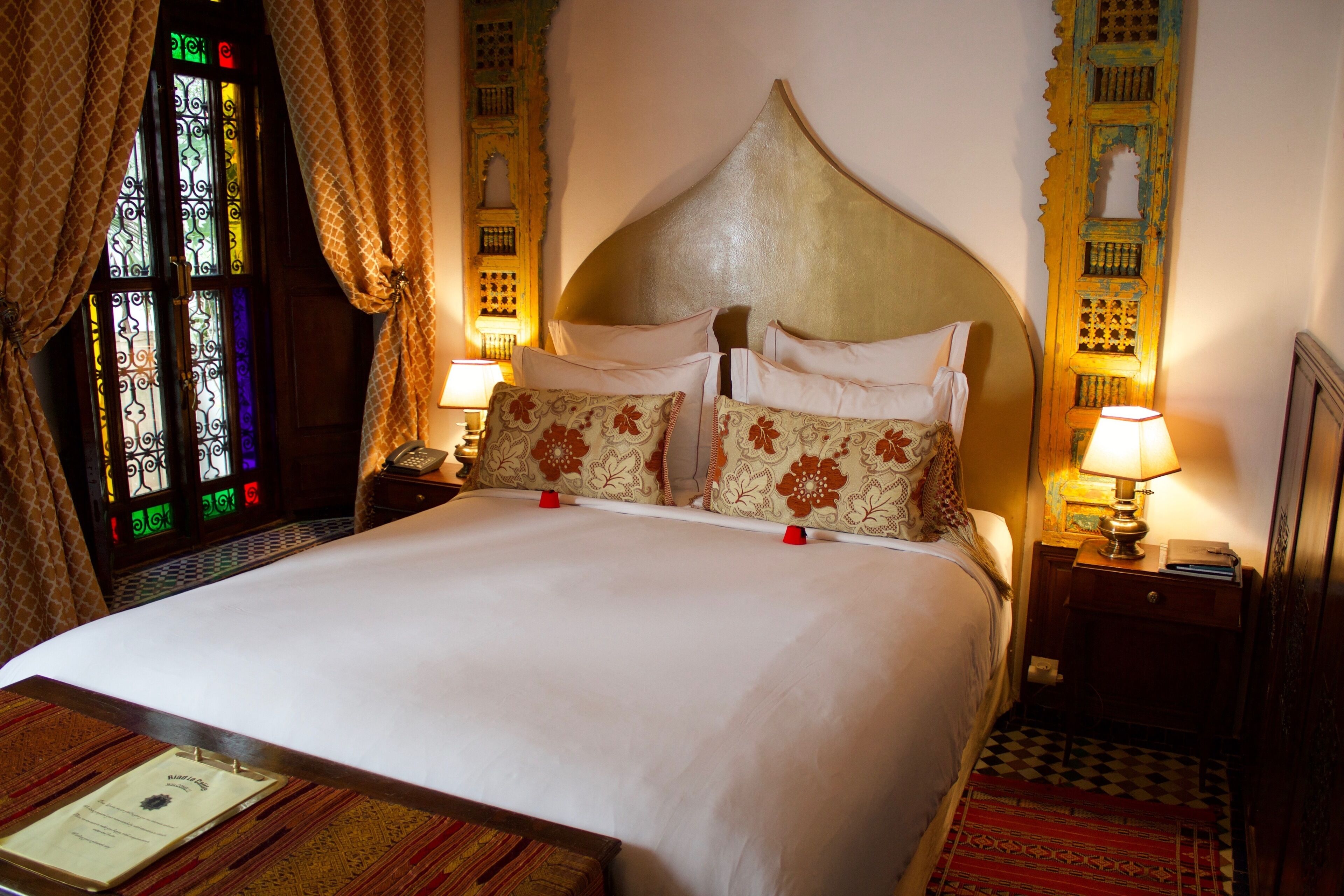 A hotel room with a large bed, a nightstand with a lamp, a window with stained glass, and a patterned carpet.