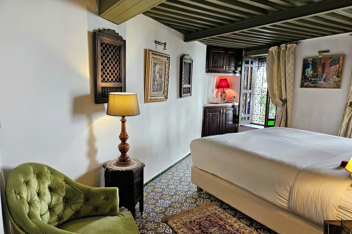 A room with a large bed, a green armchair, a wooden table with a lamp, and patterned rugs.