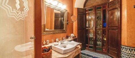 A bathroom with a double sink, a mirror with lights, a patterned tile floor, and a wooden door with glass panels.
