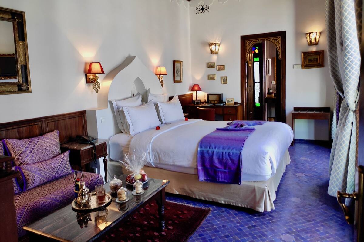 A hotel room with a large bed, a wooden nightstand, a desk with a lamp, and a small table with snacks.