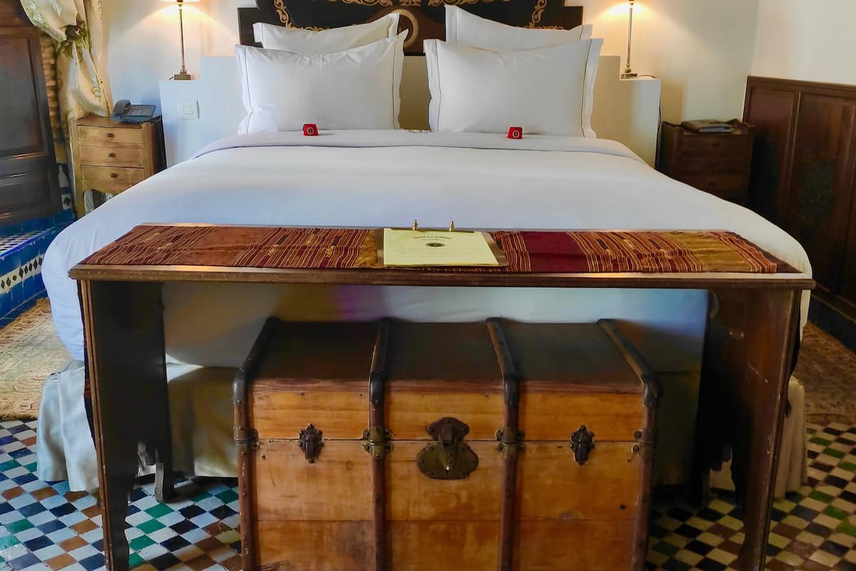 A neatly made bed with white linens, a wooden headboard with intricate carvings, two bedside lamps, a small bedside table with a phone, and a checkered floor.