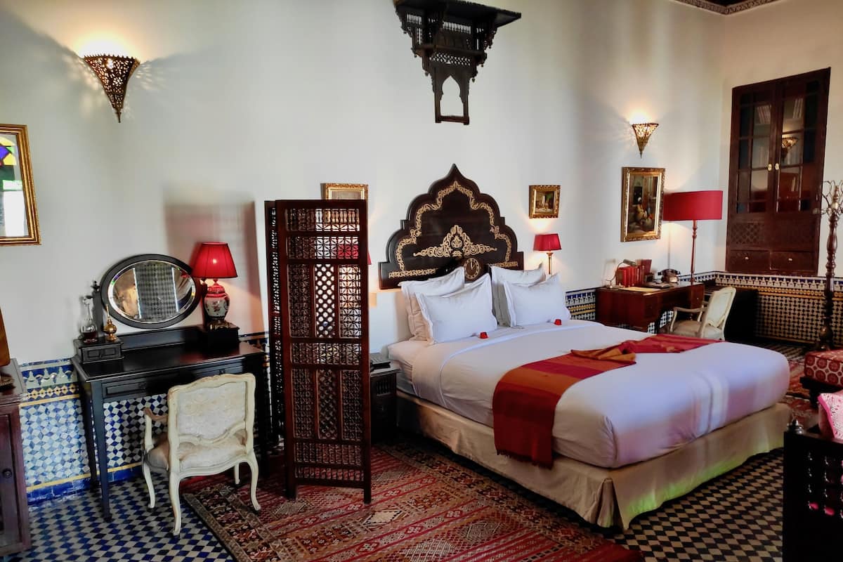 A hotel room with a large bed, a desk with a mirror, a chair, and a decorative screen.