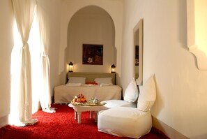 A hotel room with a red carpet, a bed with white linens, a small table with fruit and a tray, and a painting on the wall.