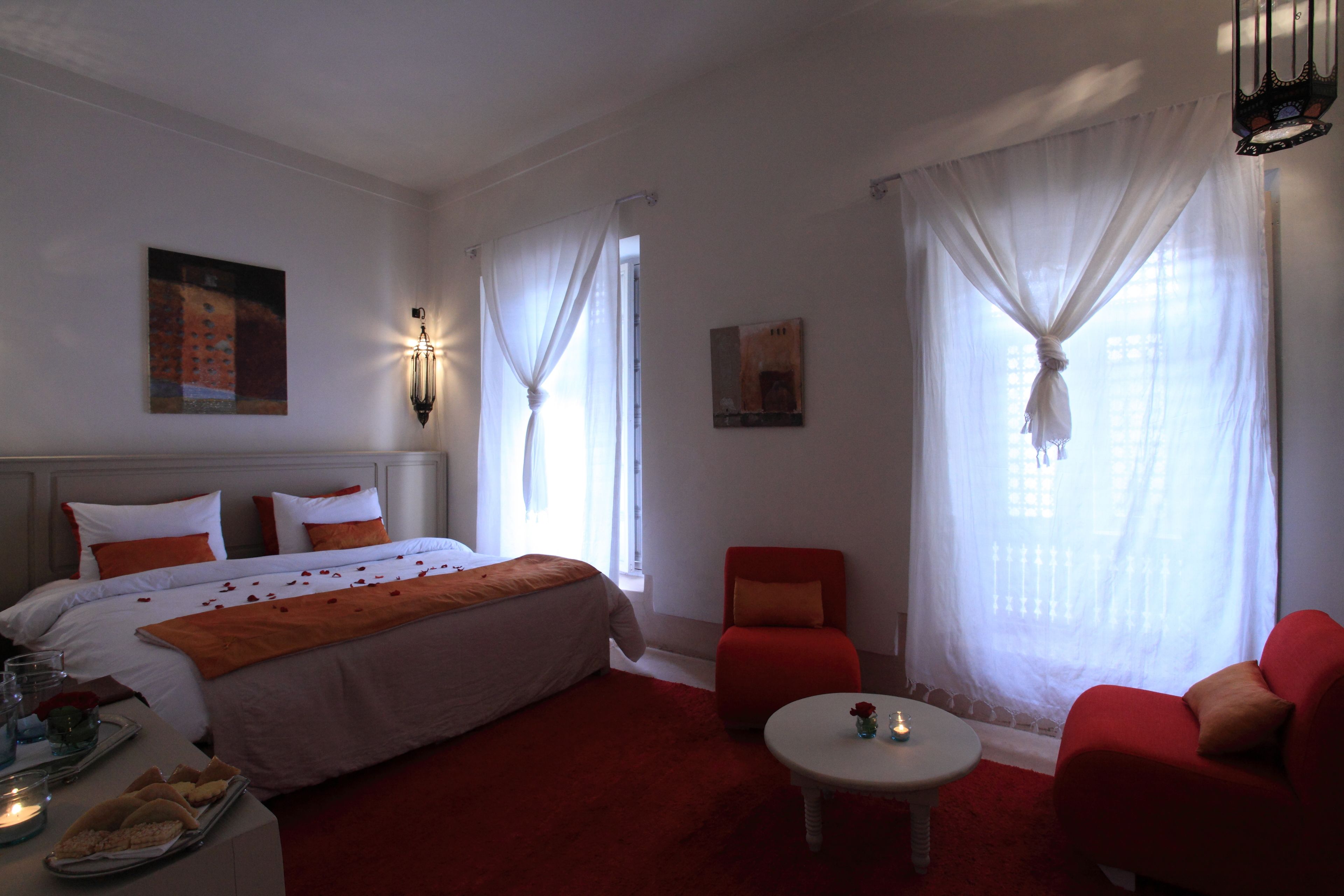 A hotel room with a large bed, a red armchair, a small table, and a window with sheer curtains.