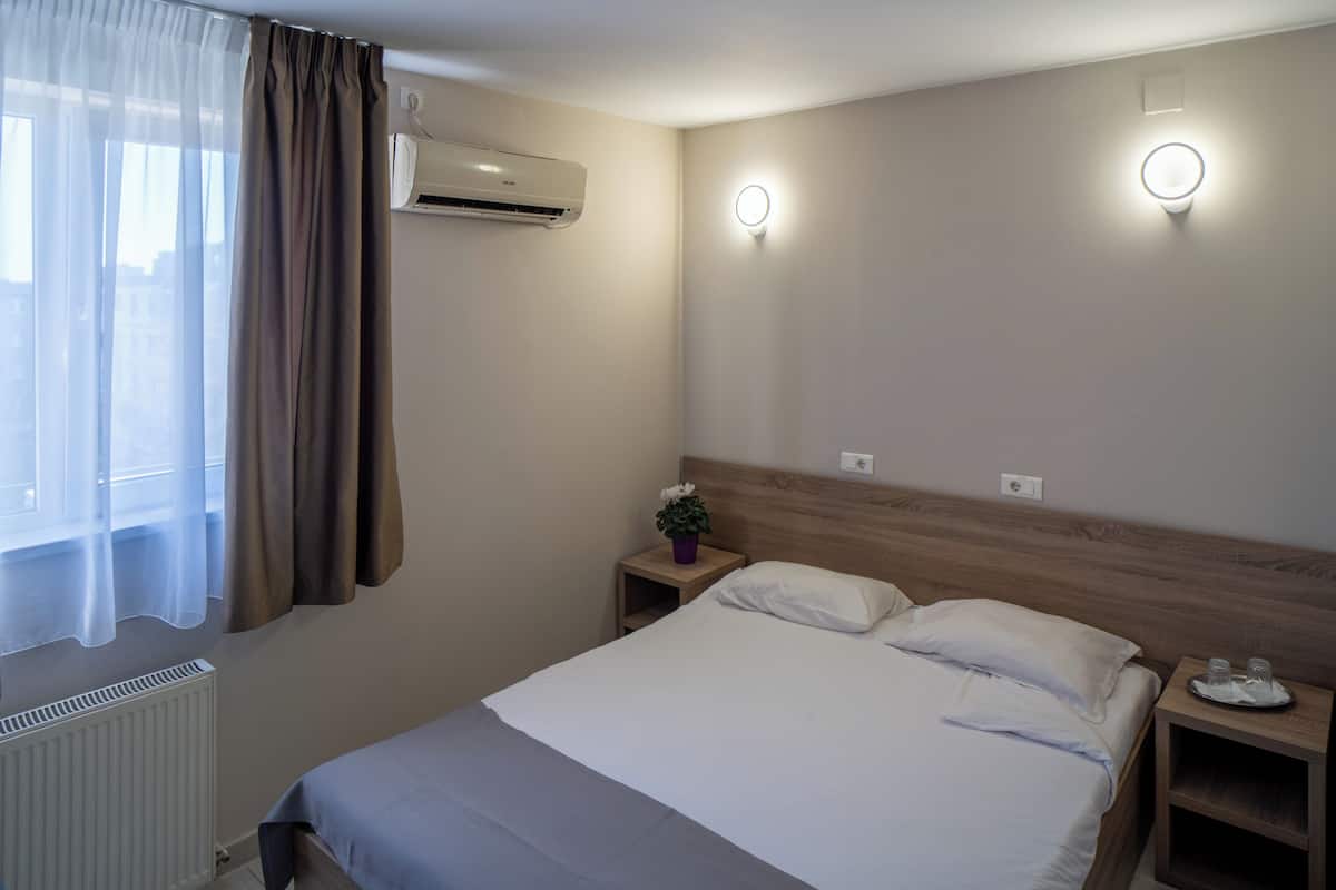 Una camera d'albergo con un letto, comodini, un tavolino da notte con una lampada, un condizionatore d'aria a muro e una finestra con tende.