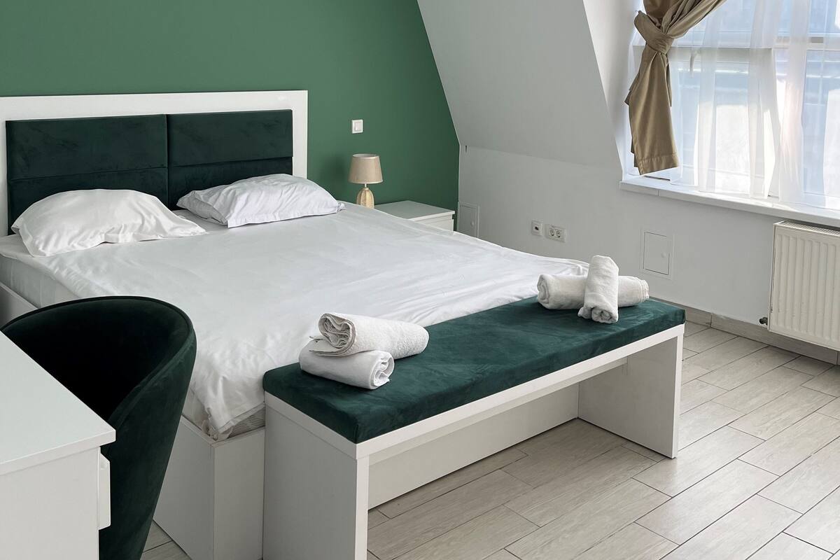 Una camera da letto moderna con una parete verde a contrasto, un letto bianco, una panca ai piedi del letto e una finestra con tende.