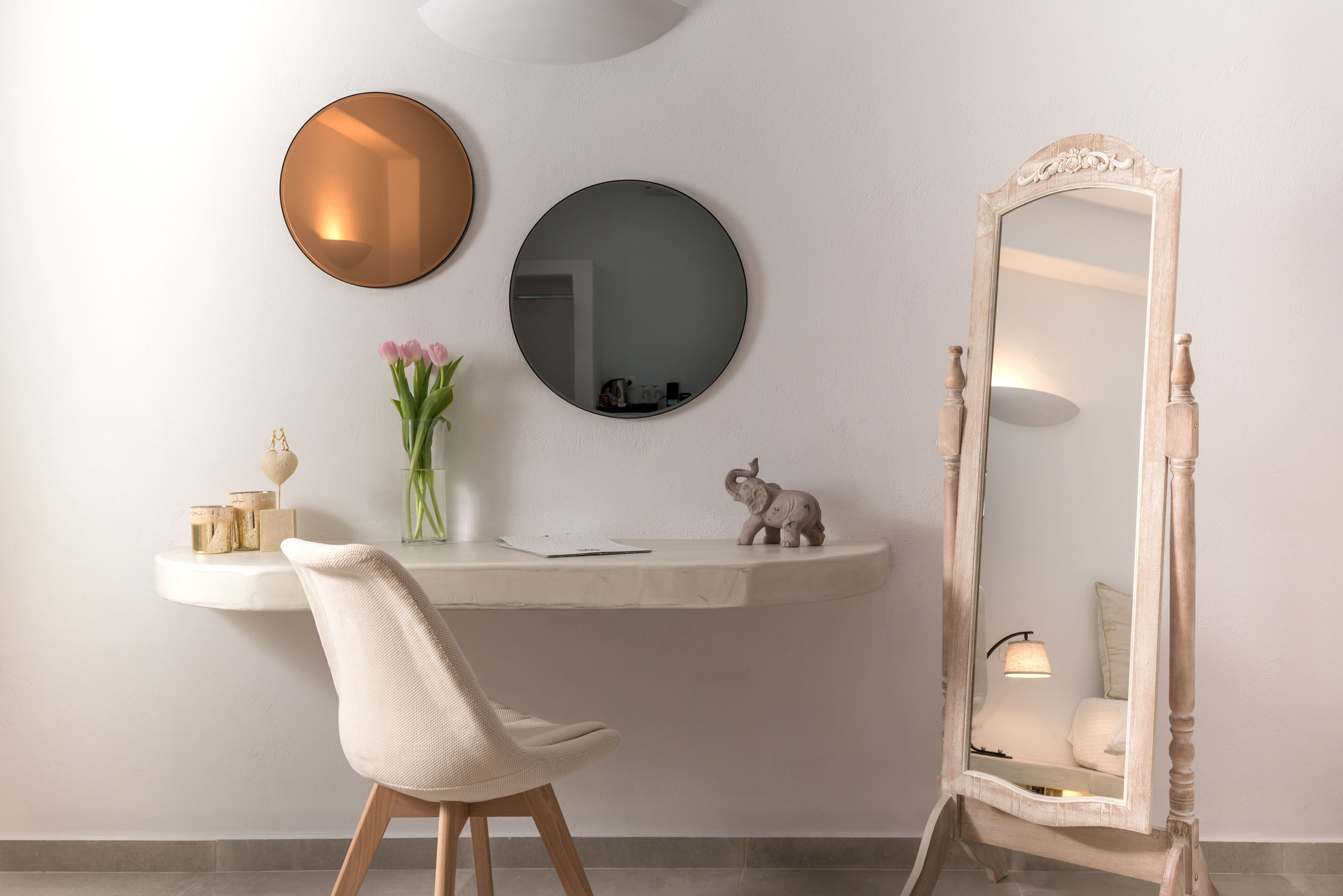 A room with a white wall, a round mirror, a round mirror, a small table with a vase of flowers, a chair, and a large mirror on a stand.