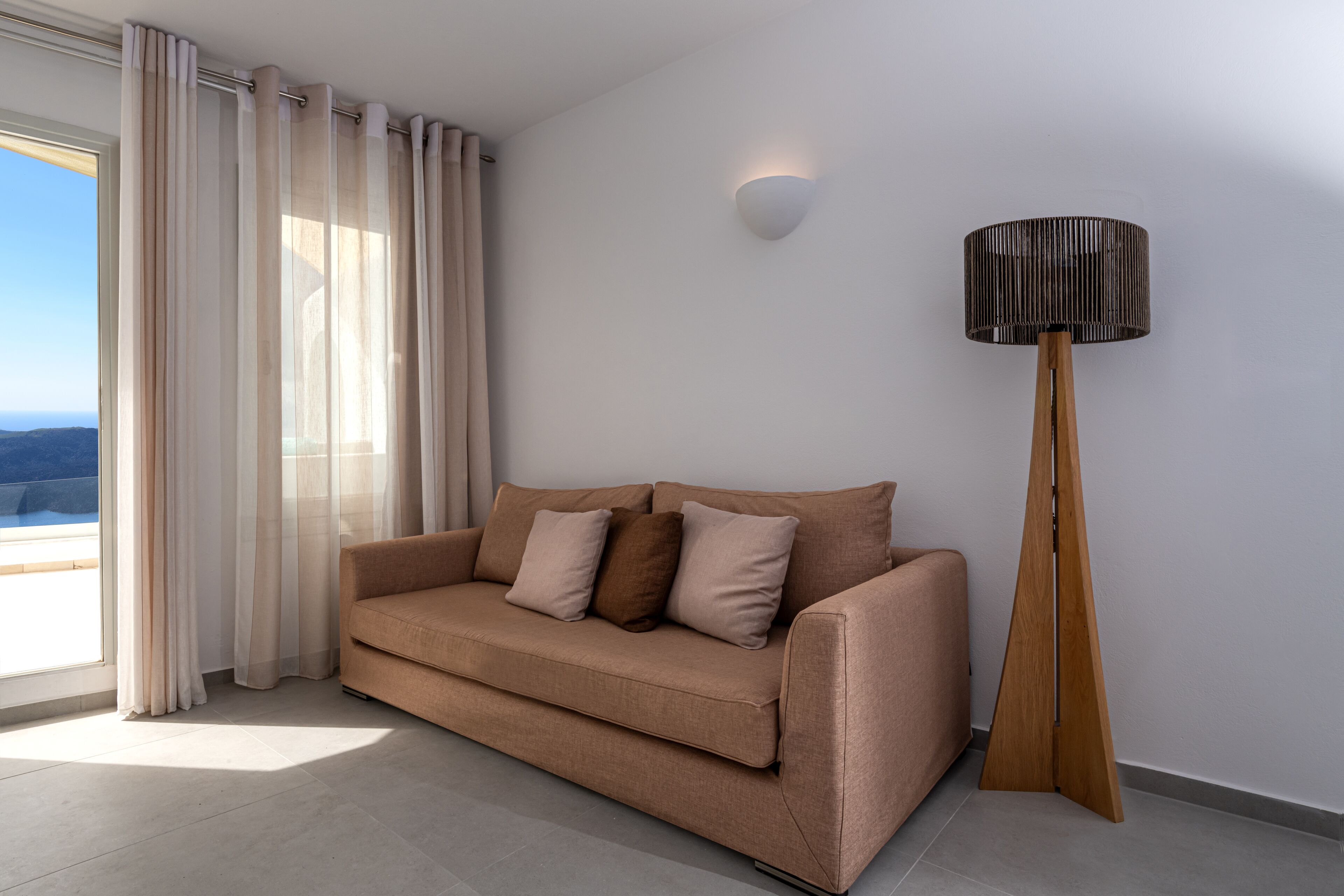 A modern living room with a beige sofa, a tall floor lamp, and a view through a large window.