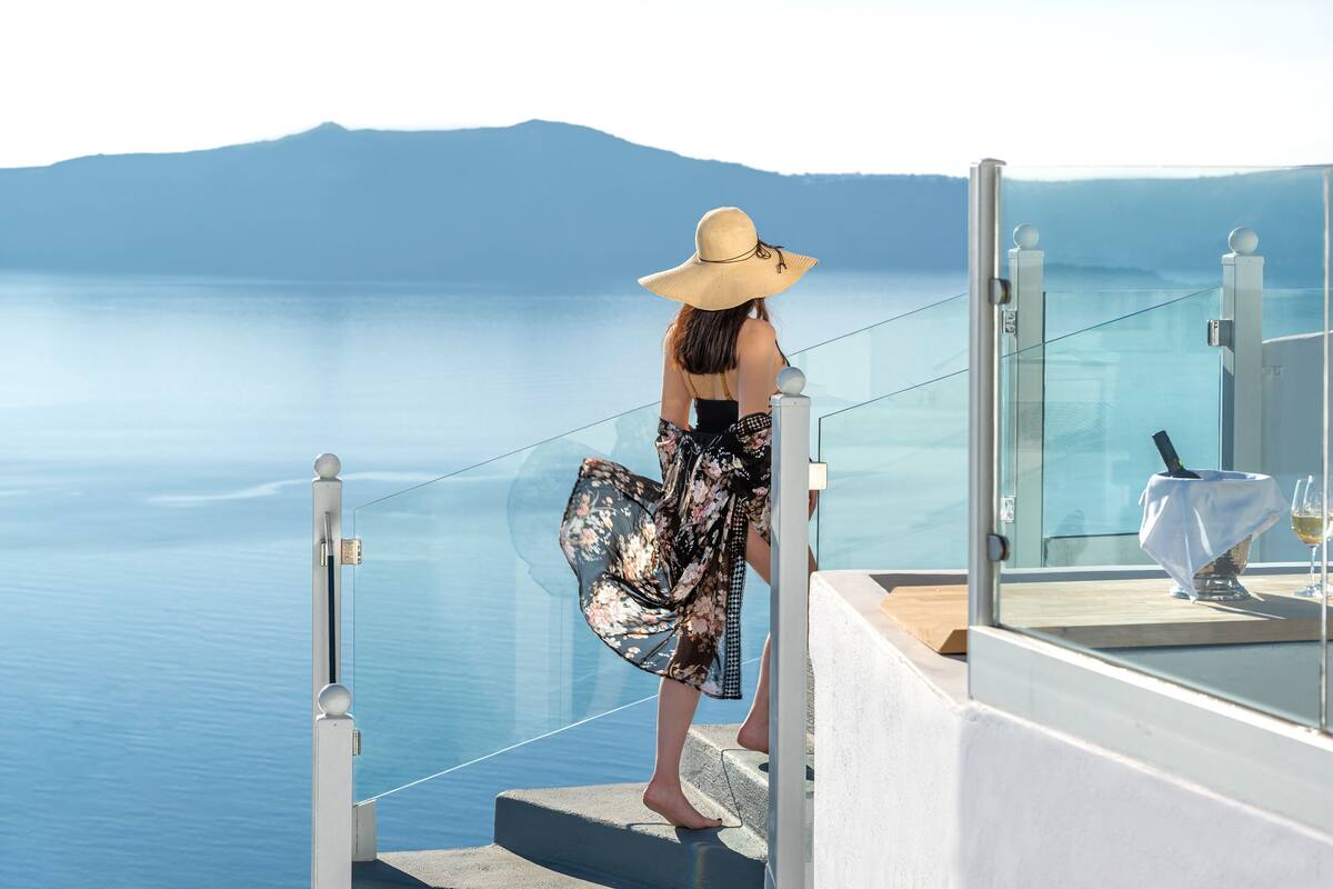 Une personne se tient sur un balcon, d’où l’on aperçoit un plan d’eau, avec des montagnes en arrière-plan.