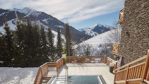 Une piscine avec une personne à l'intérieur, entourée de montagnes enneigées et d'un bâtiment en pierre.
