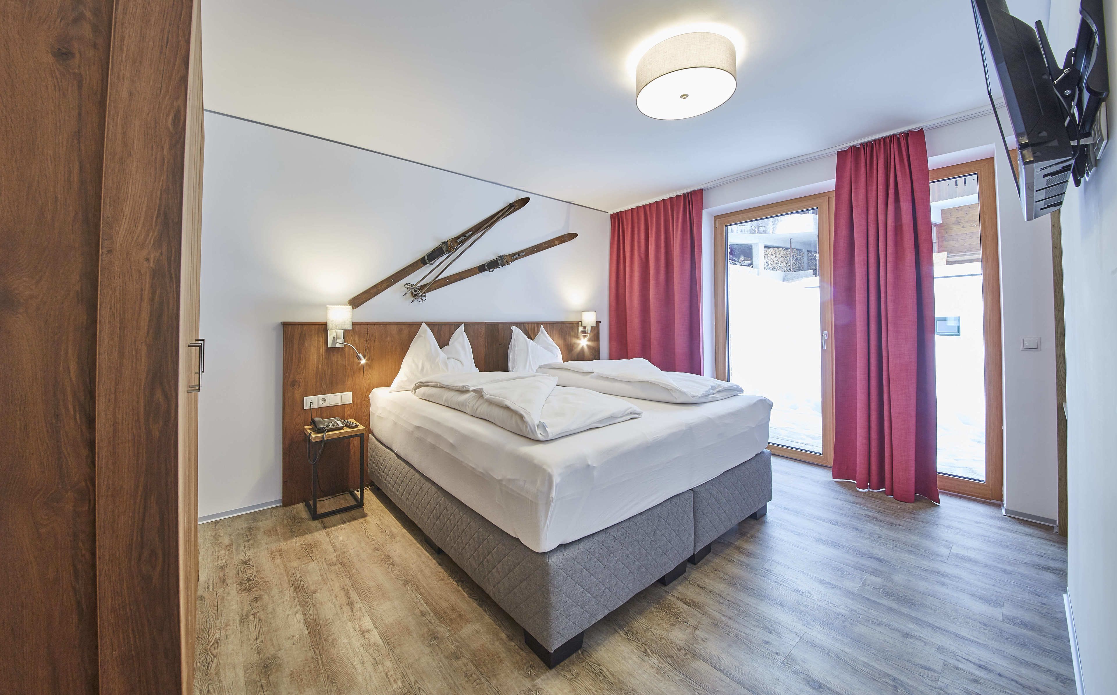 Een moderne hotelkamer met een groot bed, houten vloer, een televisie aan het plafond en een glazen schuifdeur naar een balkon.