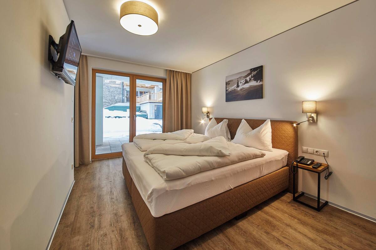 A hotel room with a bed, a TV, a bedside table with a lamp, a painting on the wall, and a view of a snowy landscape through the window.