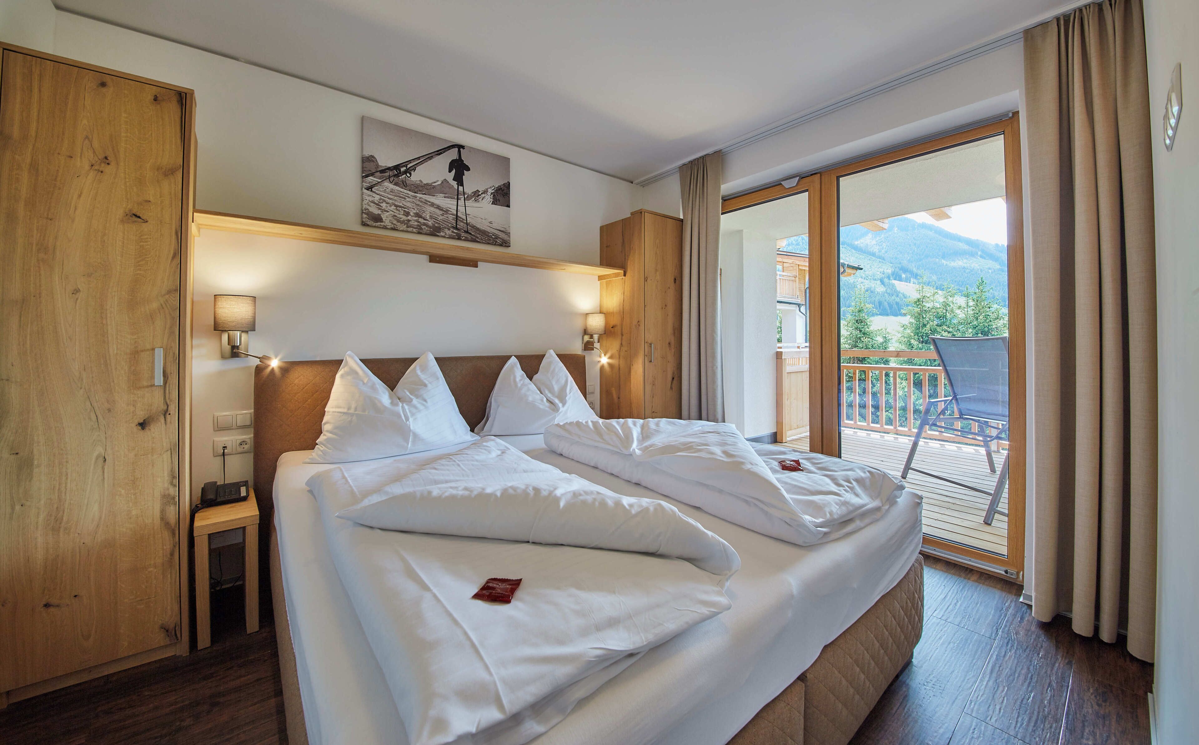 A hotel room with a large bed, a wooden headboard, a painting above the bed, a balcony with a chair, and a view of the mountains.