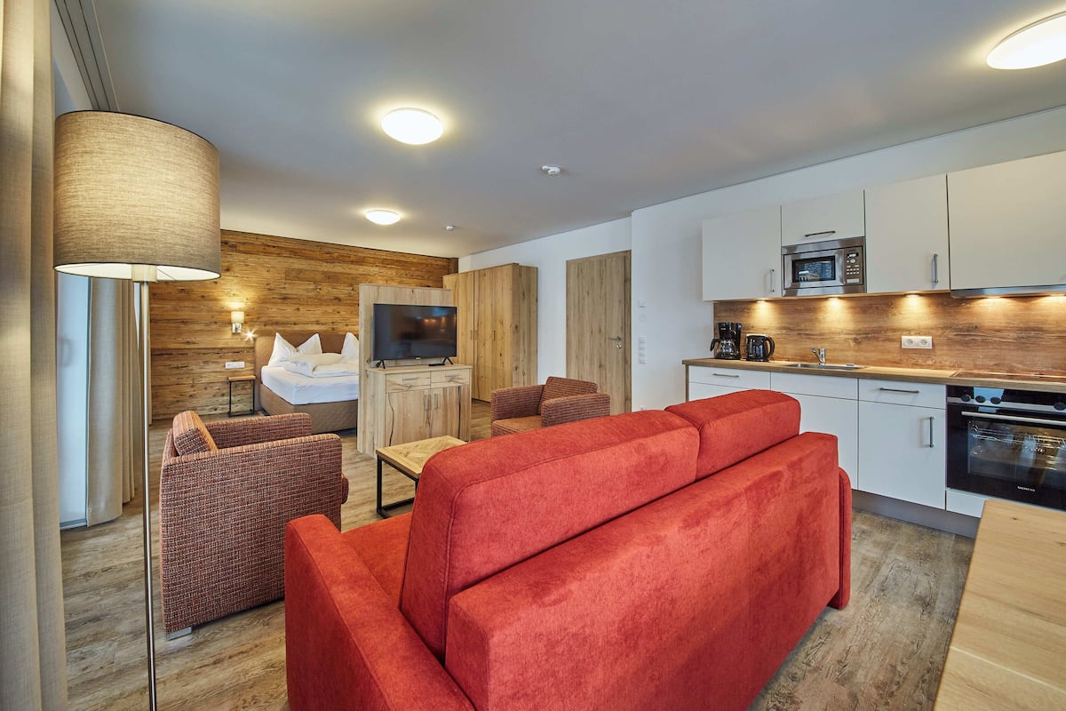 A modern hotel room with a wooden accent wall, a large bed, a TV, a sofa, a dining table, and a kitchen area.