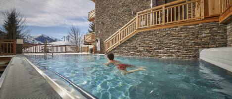 Una piscina con una persona nadando, rodeada de arquitectura de piedra y madera, y montañas al fondo.